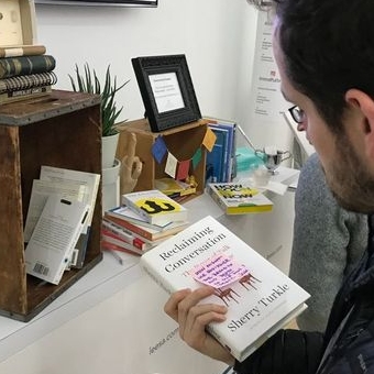 A photo of a man looking at a book near a small bookshelf.