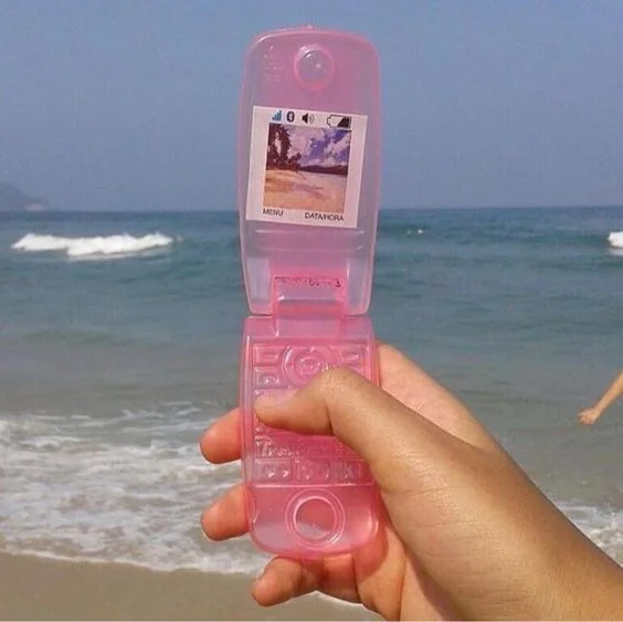 A photo of a person holding a fake plastic phone with a sticker screen with a beach picture on it. Behind the phone is a beach and ocean.