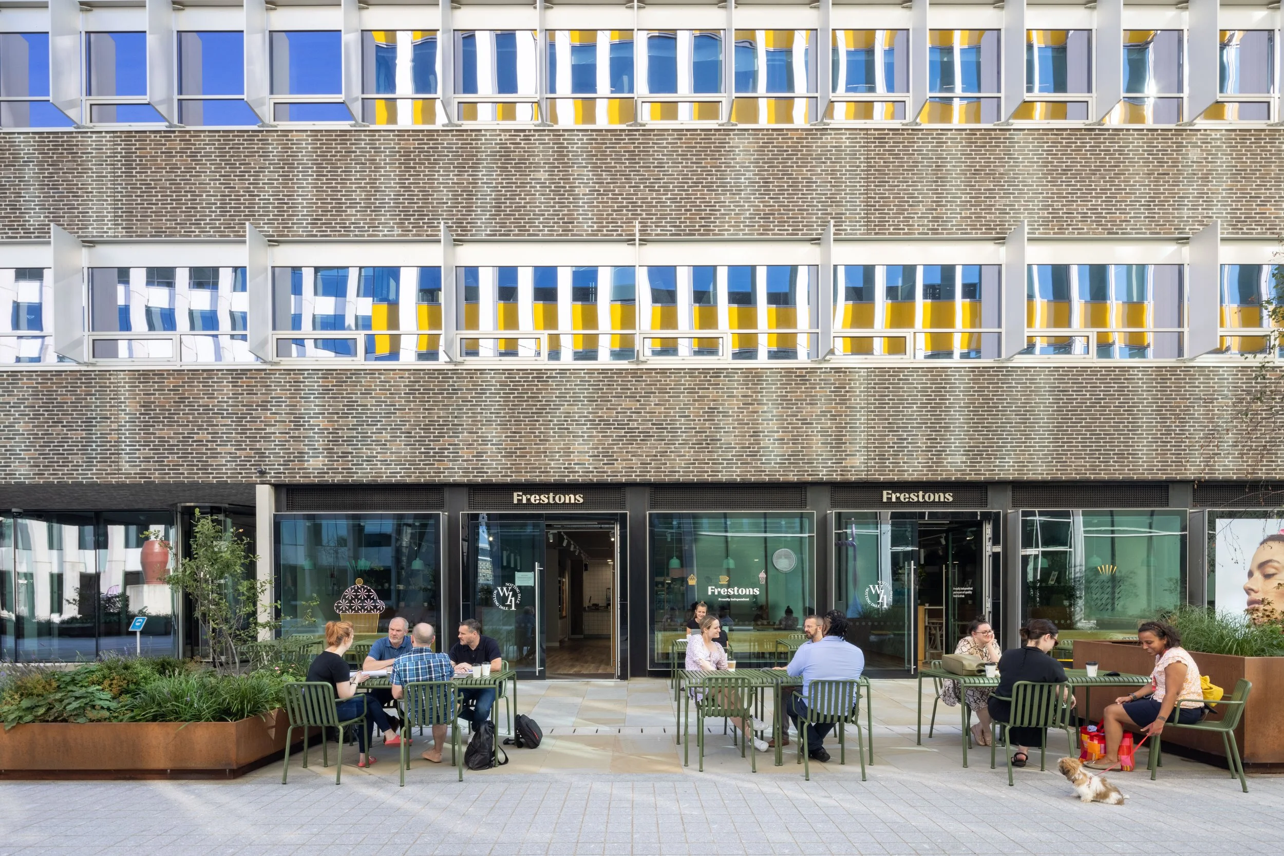 Outdoor seating with people at tables outside a brick building with large windows and signage reading 'Frestons.' A small dog sits nearby on the pavement.