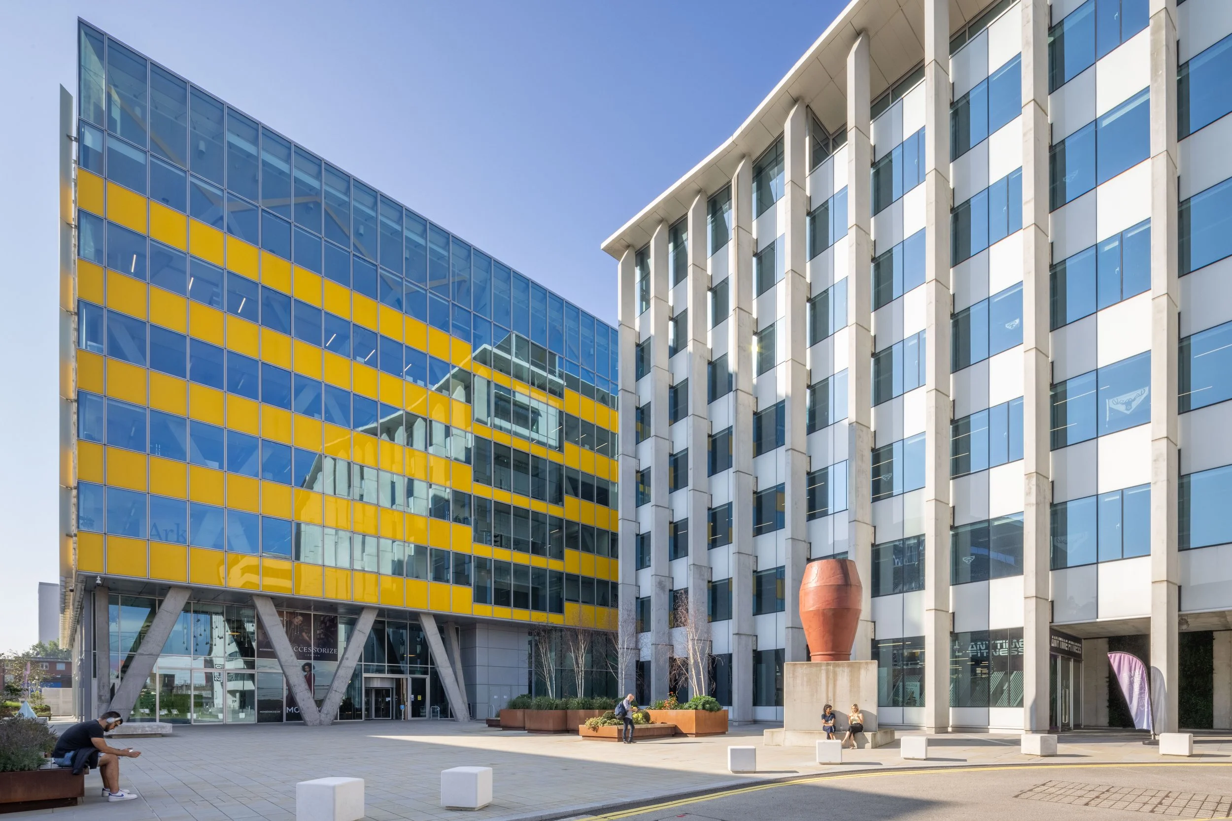 Modern office buildings with glass facades and yellow accents, featuring a courtyard with people sitting and a sculpture, under a clear blue sky.
