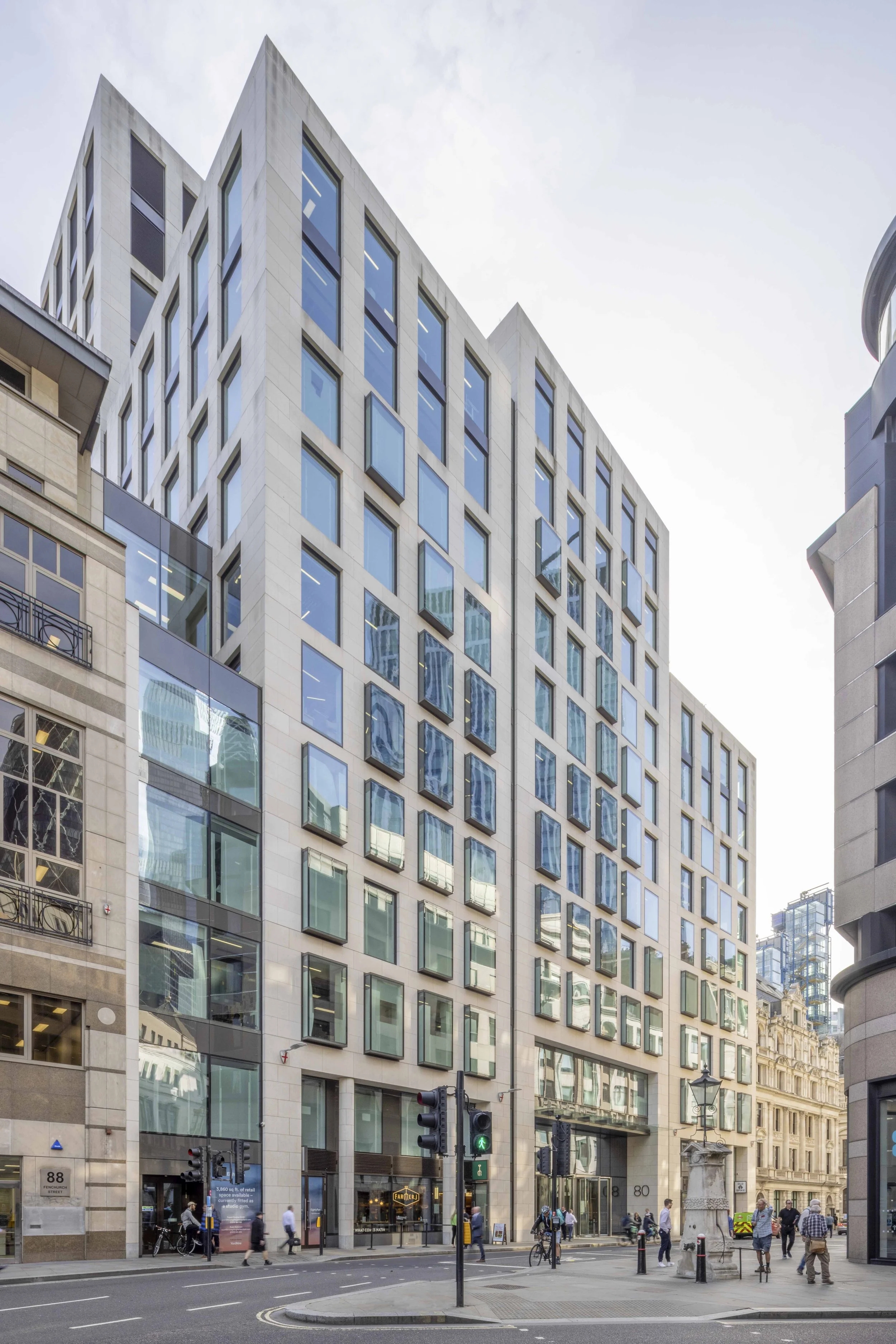 Modern glass and stone office building on a street corner with pedestrians, traffic lights, and surrounding city architecture.