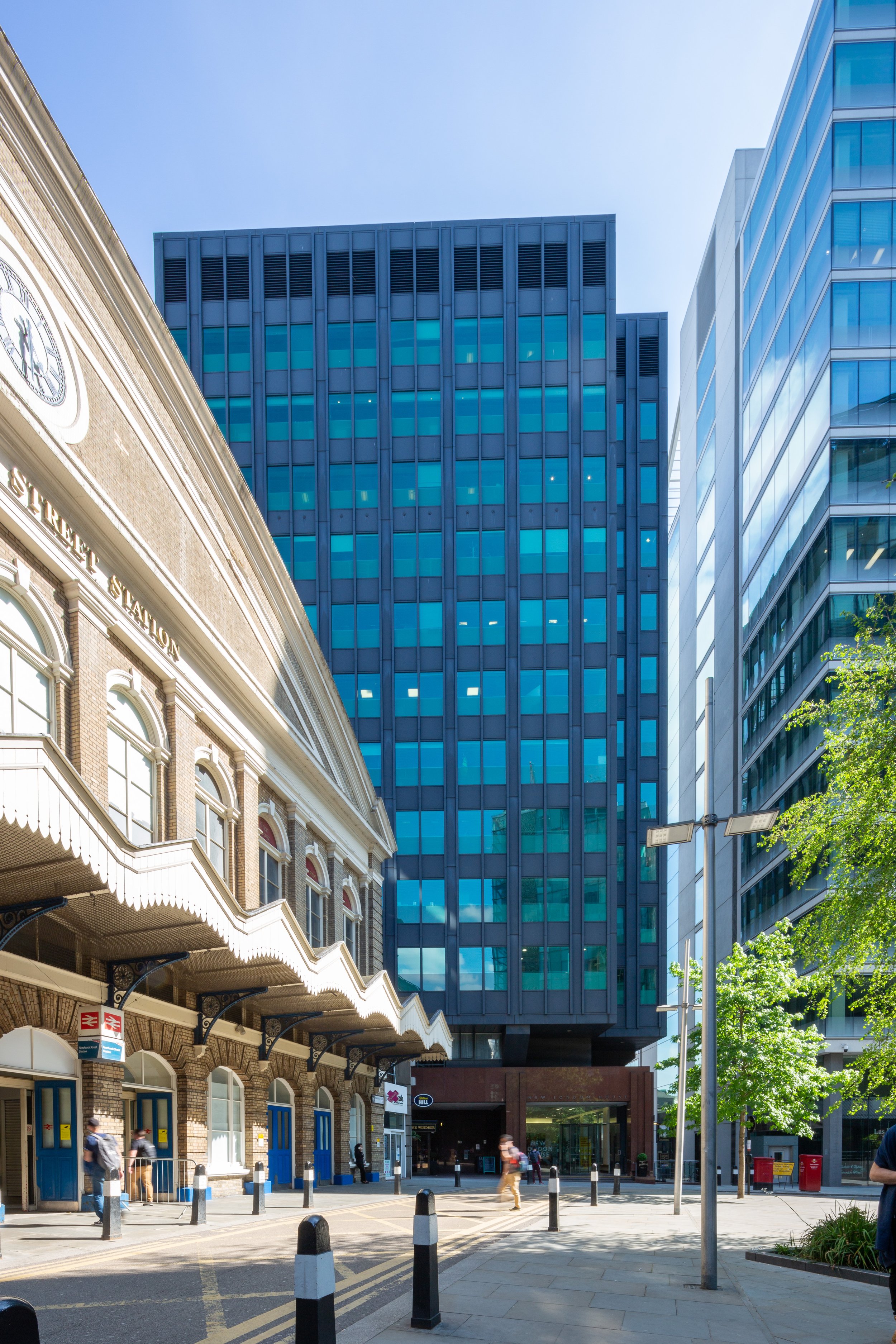 Modern and historic architectural buildings, including a brick station building and a high-rise glass office tower, with pedestrians walking on the pavement.