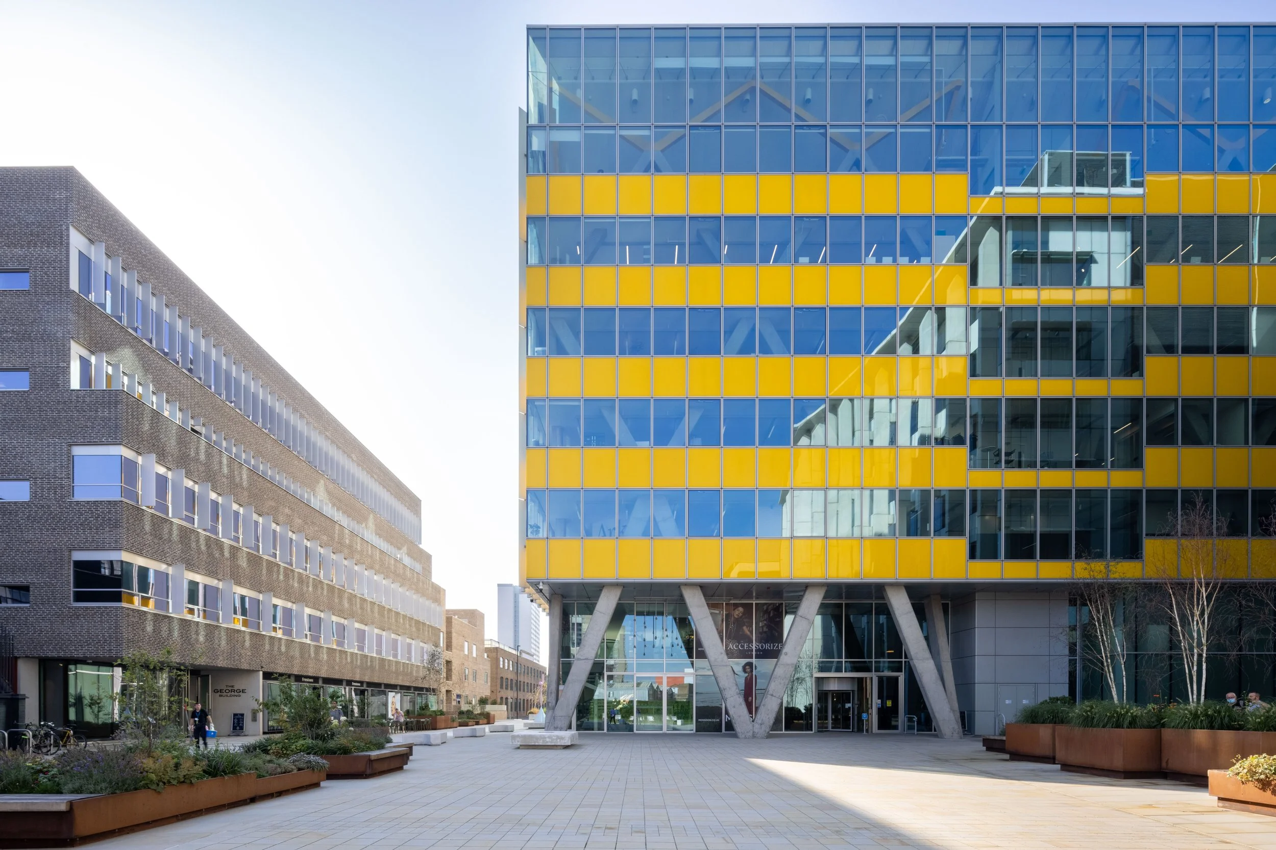 Modern office buildings with contrasting architectural designs, featuring glass facades and geometric patterns, situated in an urban setting with a paved pedestrian area.