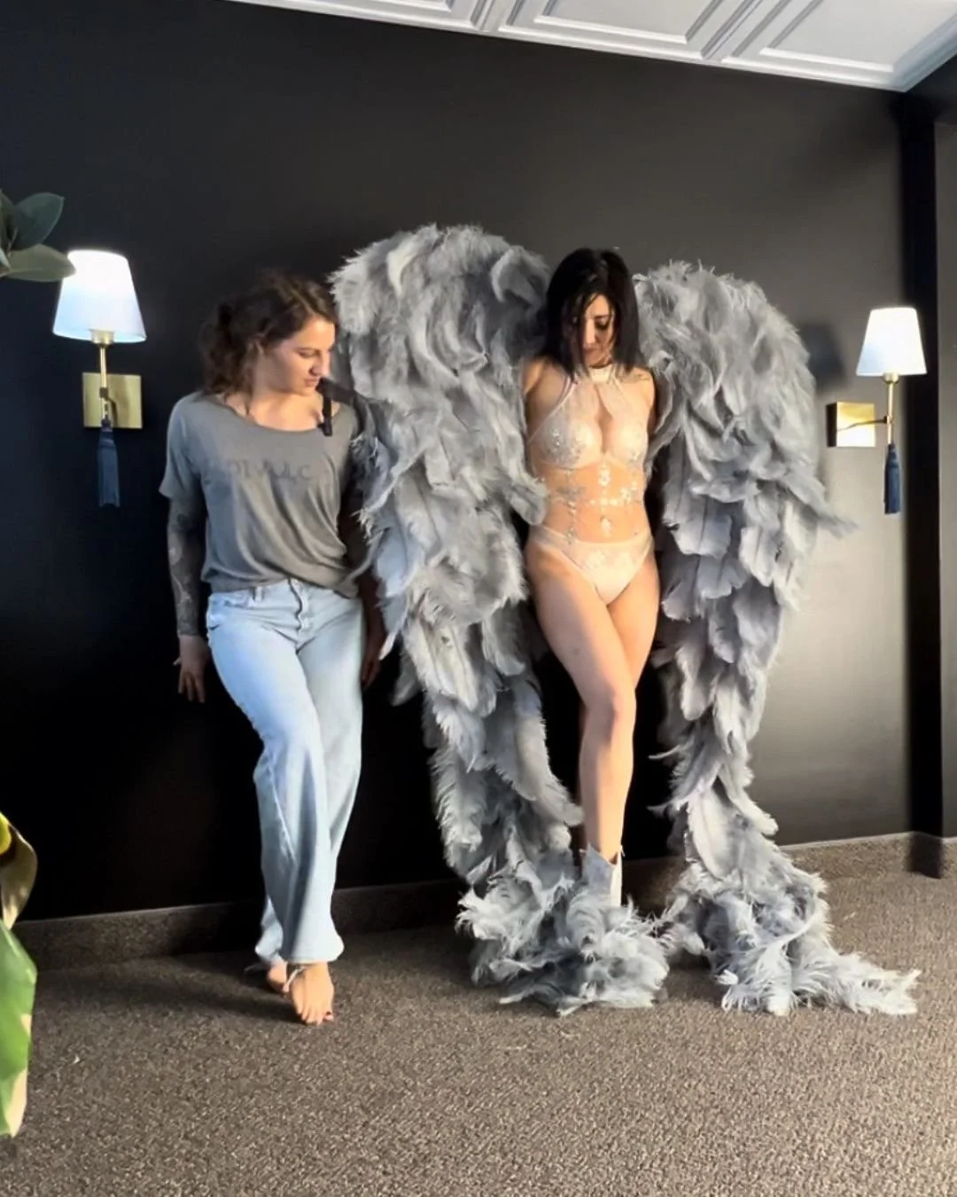 Behind-the-scenes boudoir photoshoot showing posing guidance beside a model wearing large gray feathered angel wings.