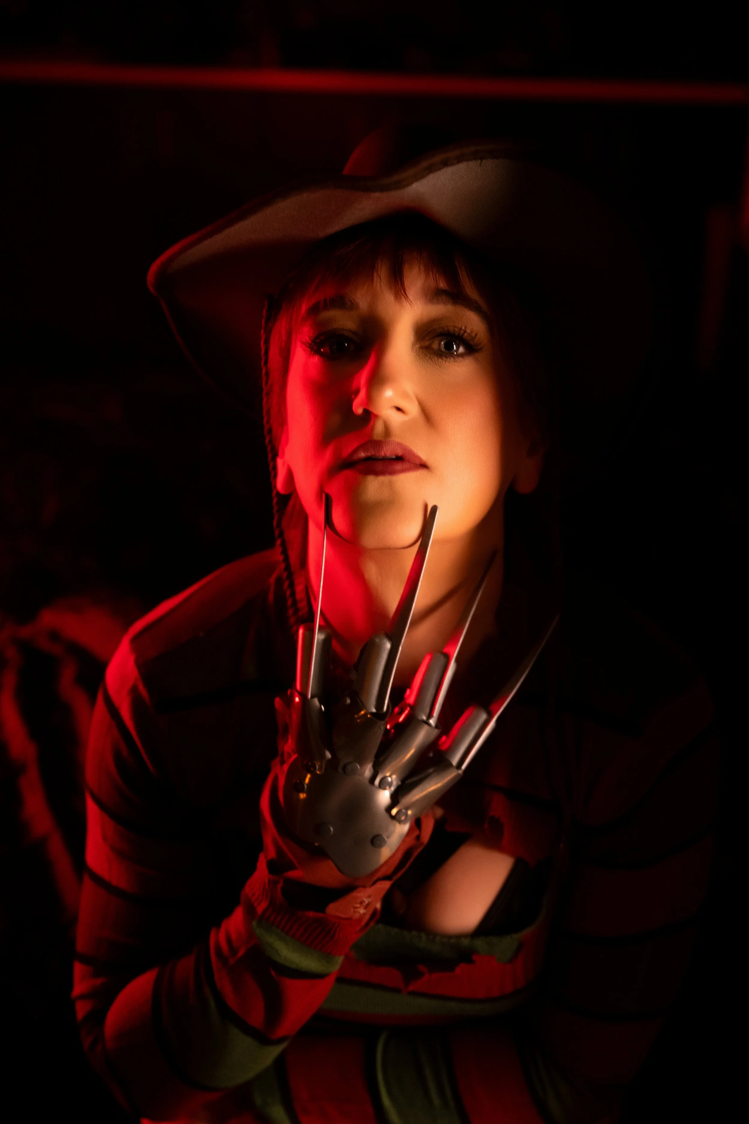 Woman in a Freddy Krueger-inspired boudoir look poses under red lighting wearing a fedora, striped sweater, and metal claw glove.