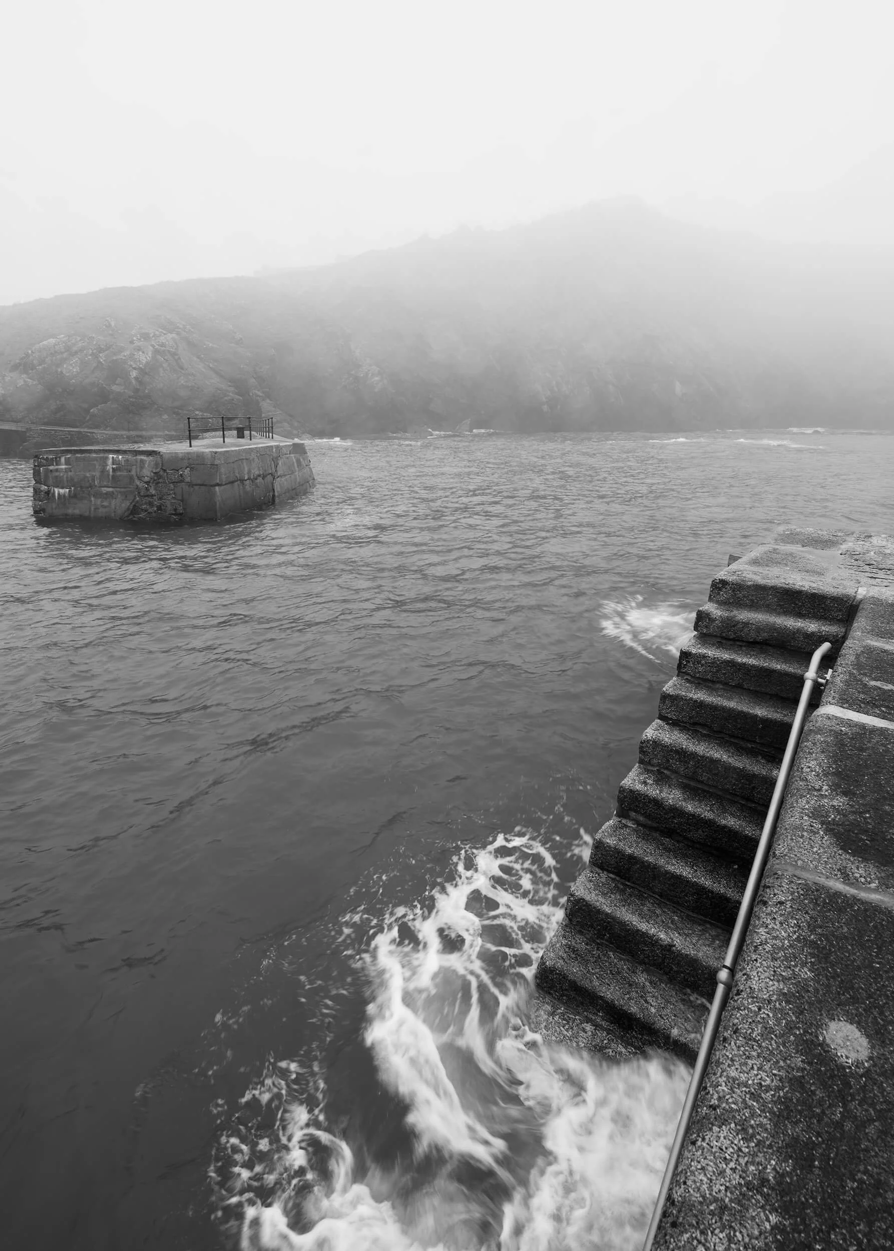 An evocative black and white photograph captures the atmospheric Mullion Harbour in Cornwall, UK, on a misty or foggy day. The monochrome palette intensely highlights the textures of the stone steps leading into the turbulent water on the right, and 