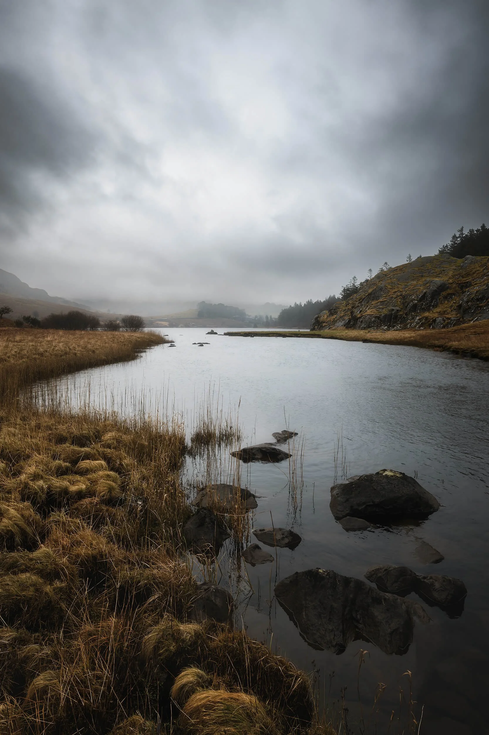 A quiet stretch of water in North Wales under a heavy sky. Wind-bent grasses line the shoreline while dark rocks emerge through the still surface, drawing the eye gently into the misty distance.

Photographed on a grey, rain-heavy afternoon, the subd