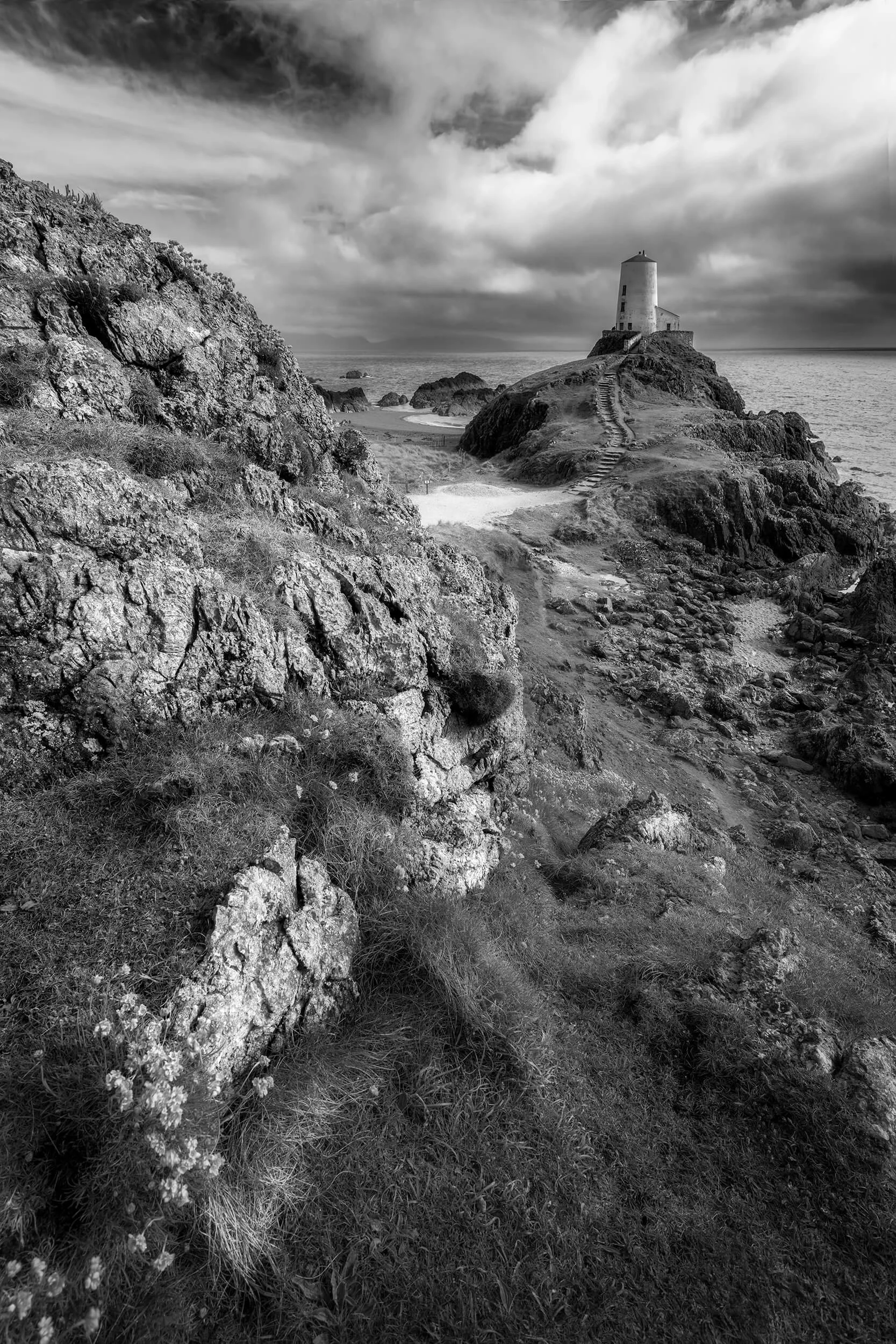 A dramatic black and white landscape photograph captures the iconic Twr Mawr Lighthouse standing proudly on the rocky island of Ynys Llanddwyn, Anglesey, North Wales. The monochrome palette accentuates the rugged textures of the foreground rocks and 