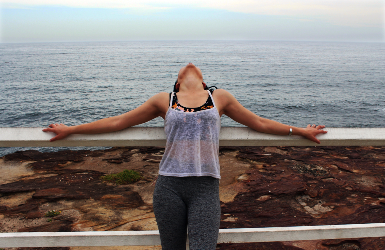 [image description: Ria in activewear leaning against a fence with the sea in the background. Her head is leaning so far back that you can only see her chin and neck, and her arms are stretched across the fence]
