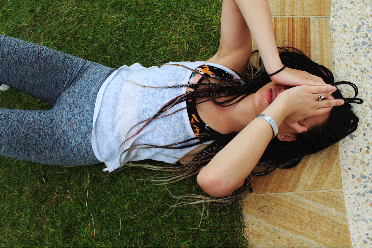 [image description: Ria in activewear lying on grass and her head is on a sandstone pavement. She is covering her face with her hands.]