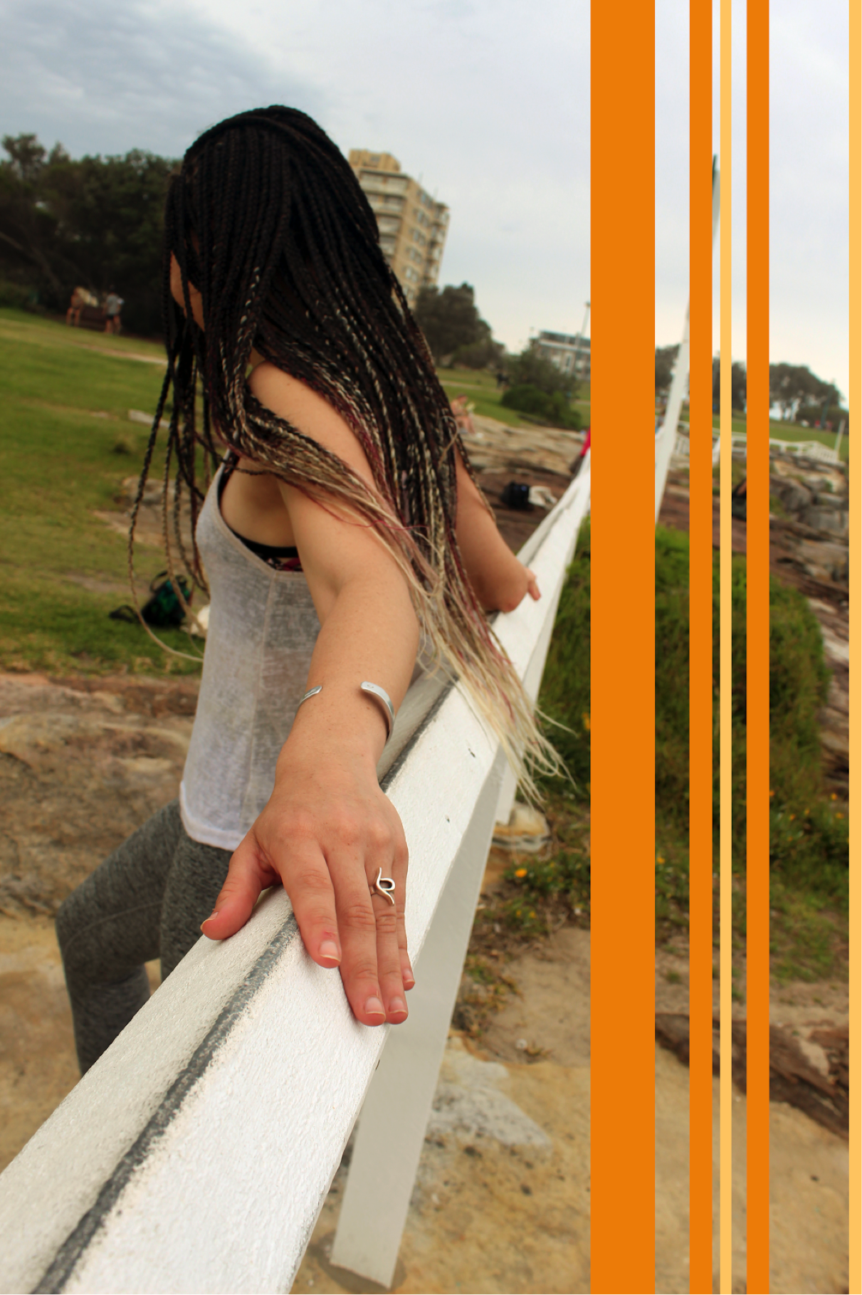 [image description: image of Ria leaning on a white fence taken from the side. You can clearly see a ring on her left hand, which has the eating disorder recovery symbol. She has brown braids which are magenta and blonde at the tips. There are orang…