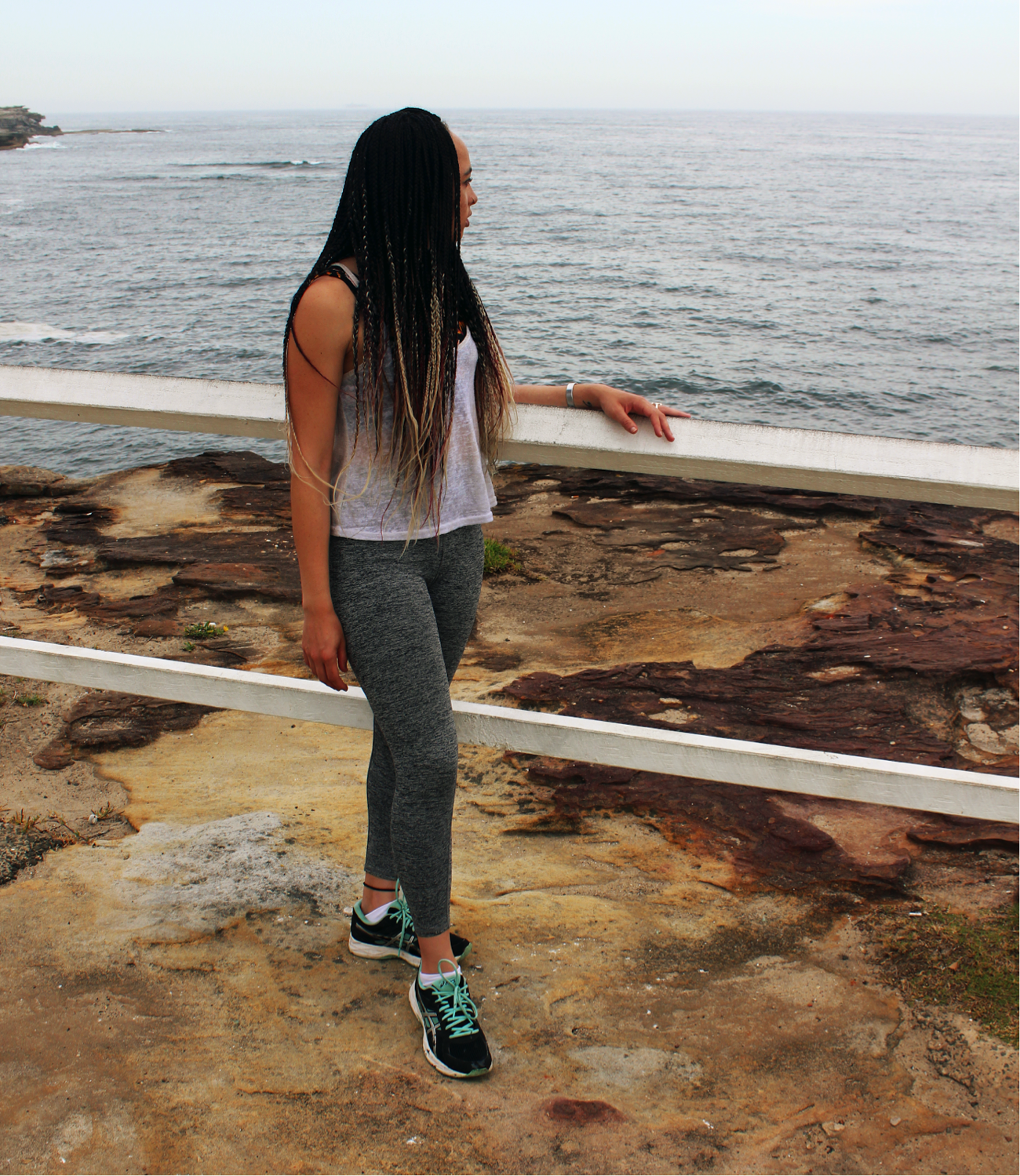[image description: Ria in activewear standing on a rock and looking at the ocean over her left shoulder. Her left arm is leaning on a white fence and her right arm is by her side.]