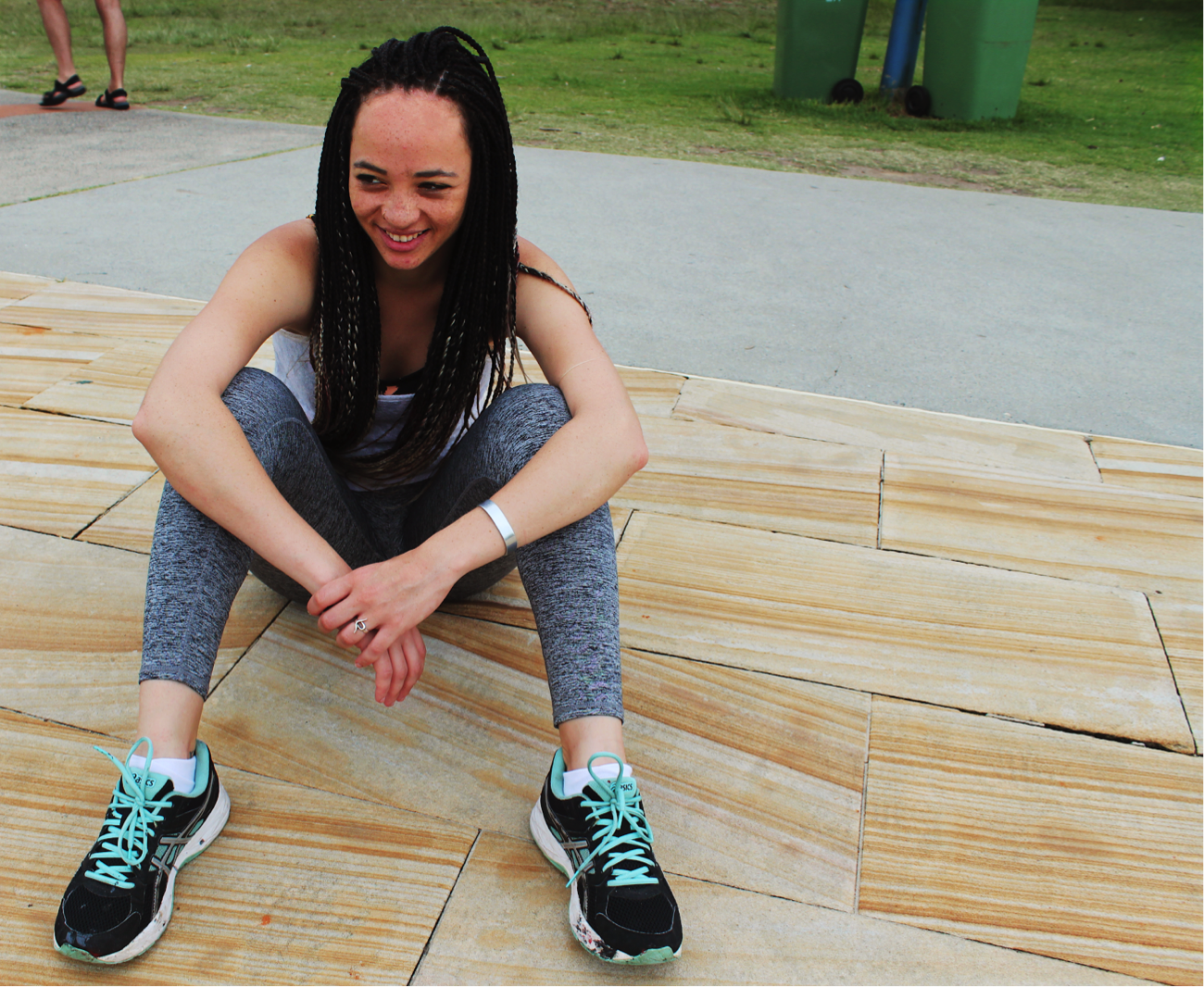 [image description: Ria is sitting on a sandstone pavement with her knees bent. Her arms are placed casually over her knees and she is smiling and looking away from the camera].