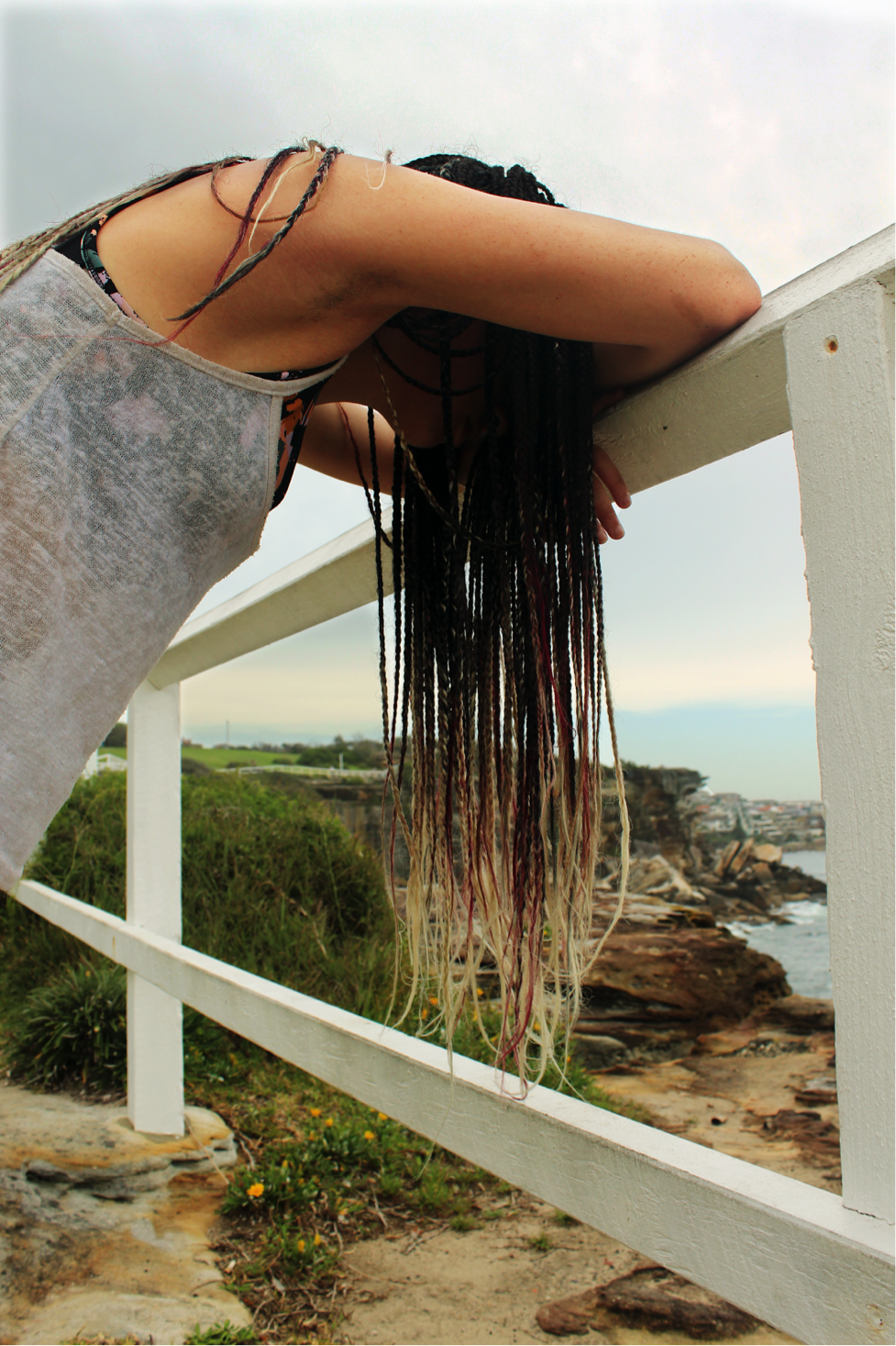 [image description: Ria is leaning her head and crossed arms on a white fence. She has long, brown braids which are magenta and blonde at the tips. You cannot see her face.]