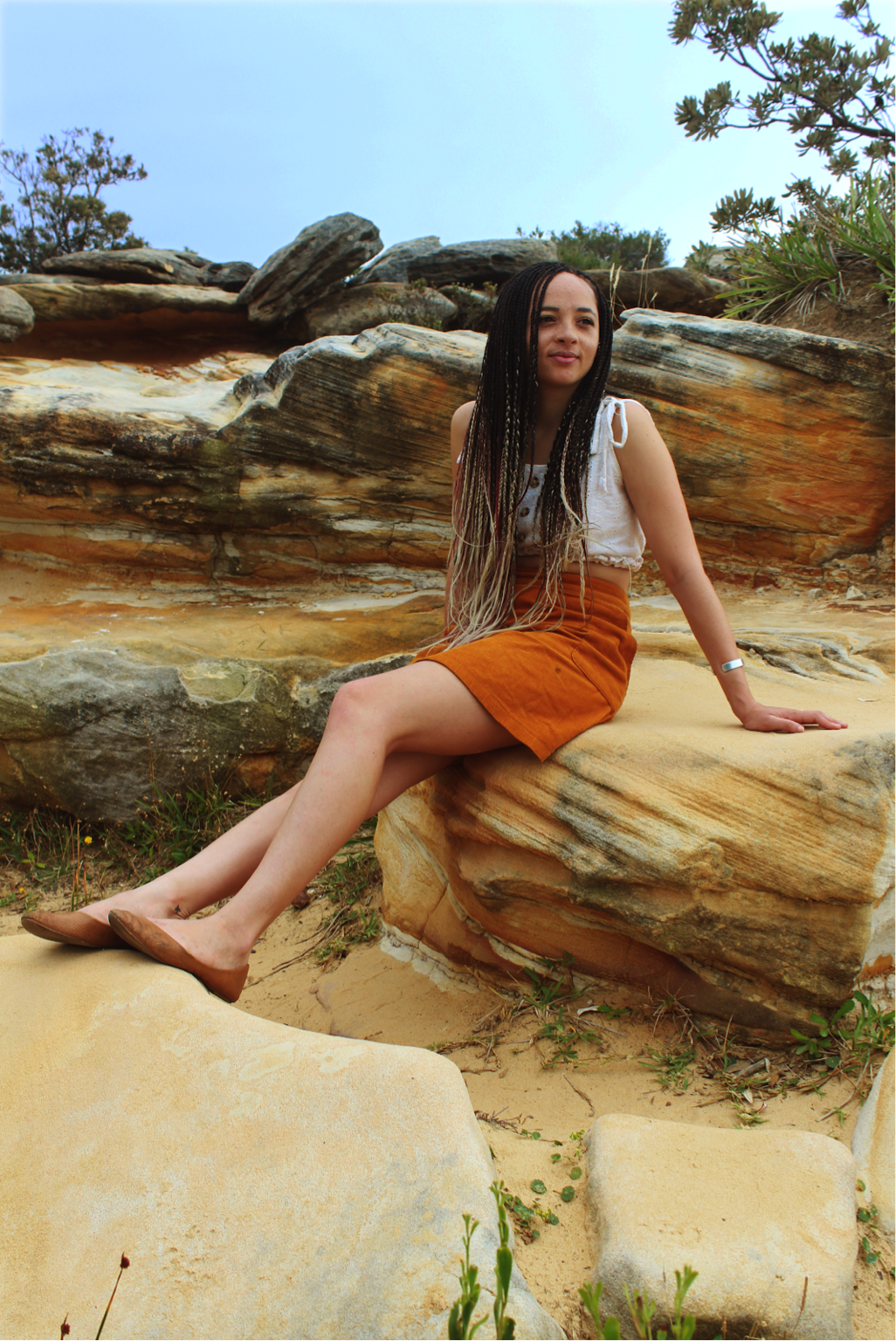 [image description: Ria is sitting on a sandstone rock, her legs are stretched out and placed on a rock in front of her. She is looking out to the distance with a relaxed expression.]