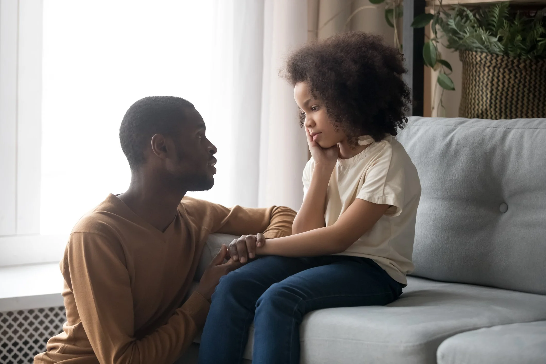 A man kneeling beside a young girl who is sitting on a sofa, both engaging in a serious conversation in a living room.