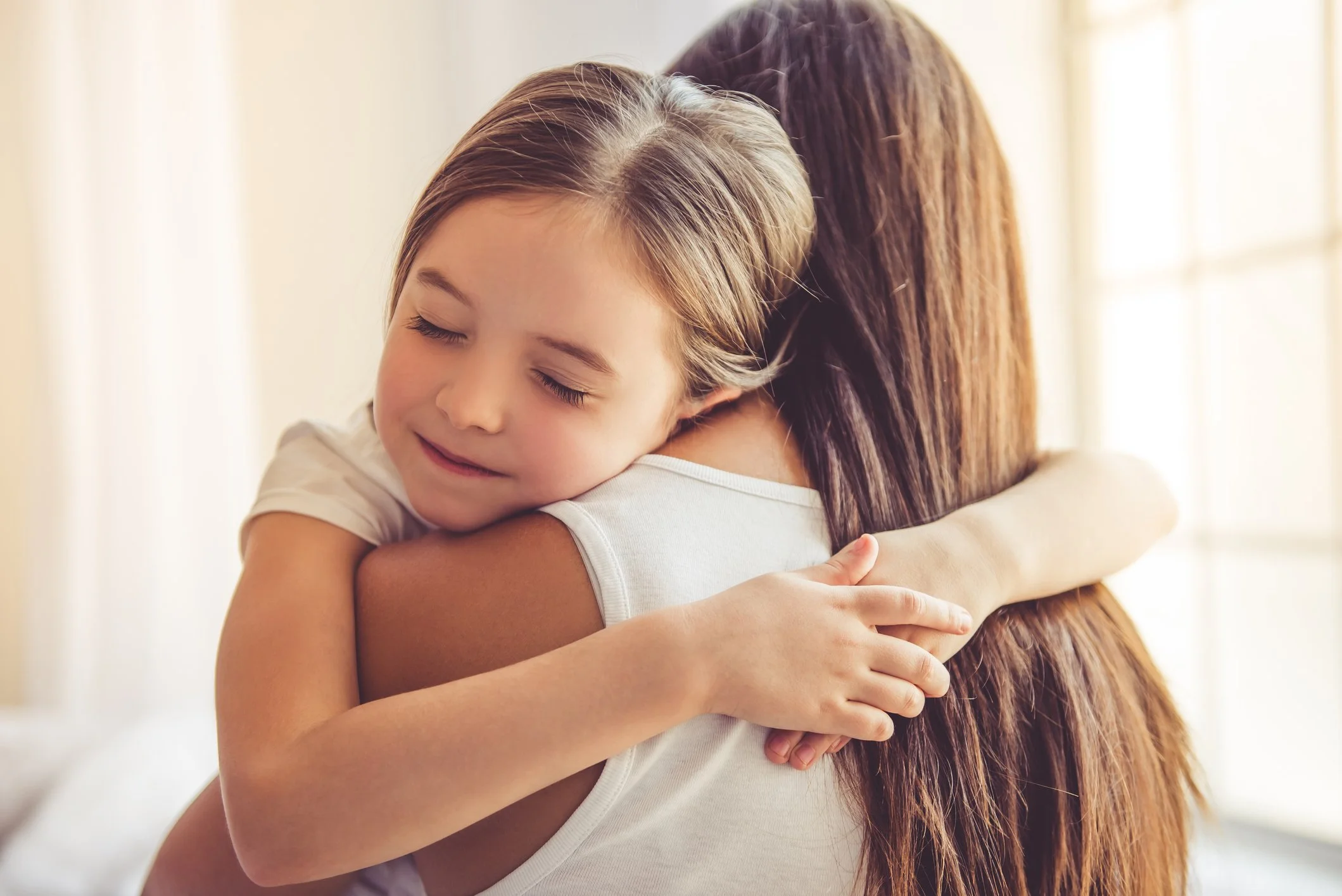 A young girl with closed eyes hugging an adult woman, likely her mother, in a warm embrace indoors.