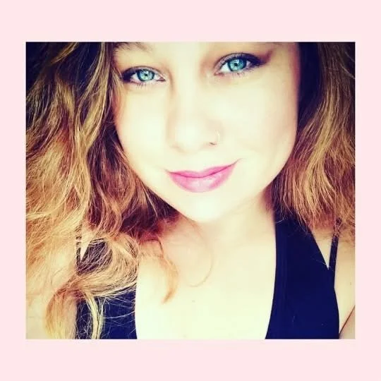 Close-up of a young woman with curly, light brown hair, blue eyes, and light makeup, smiling softly at the camera. She is wearing a black top with spaghetti straps, and the background is pink.