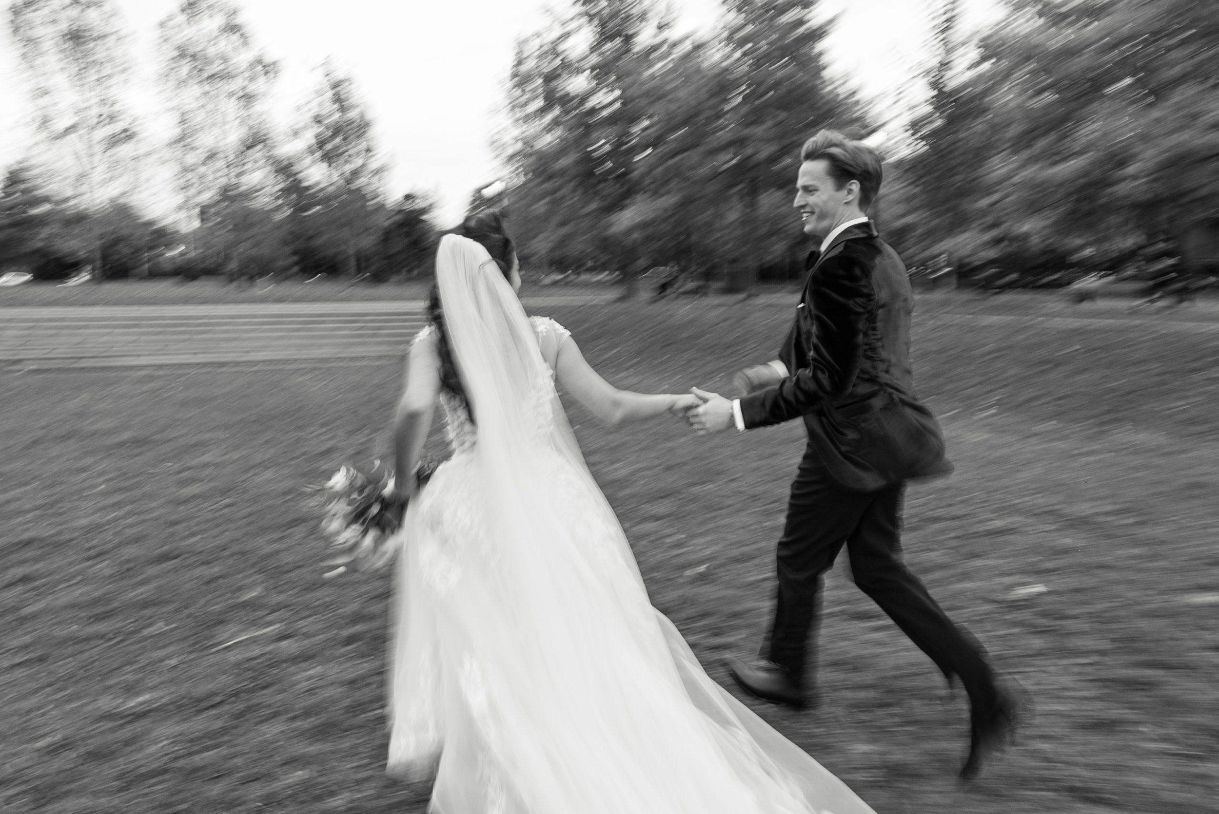 A beautiful black and white blur of the bride and groom running away together in an urban Montreal field of lush green.
