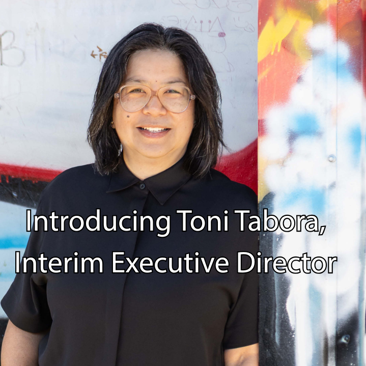 "Introducing Toni Tabora, Interim Executive Director" in white lettering over a photo of a smiling person wearing a black button up shirt and standing in front of a colorful exterior wall