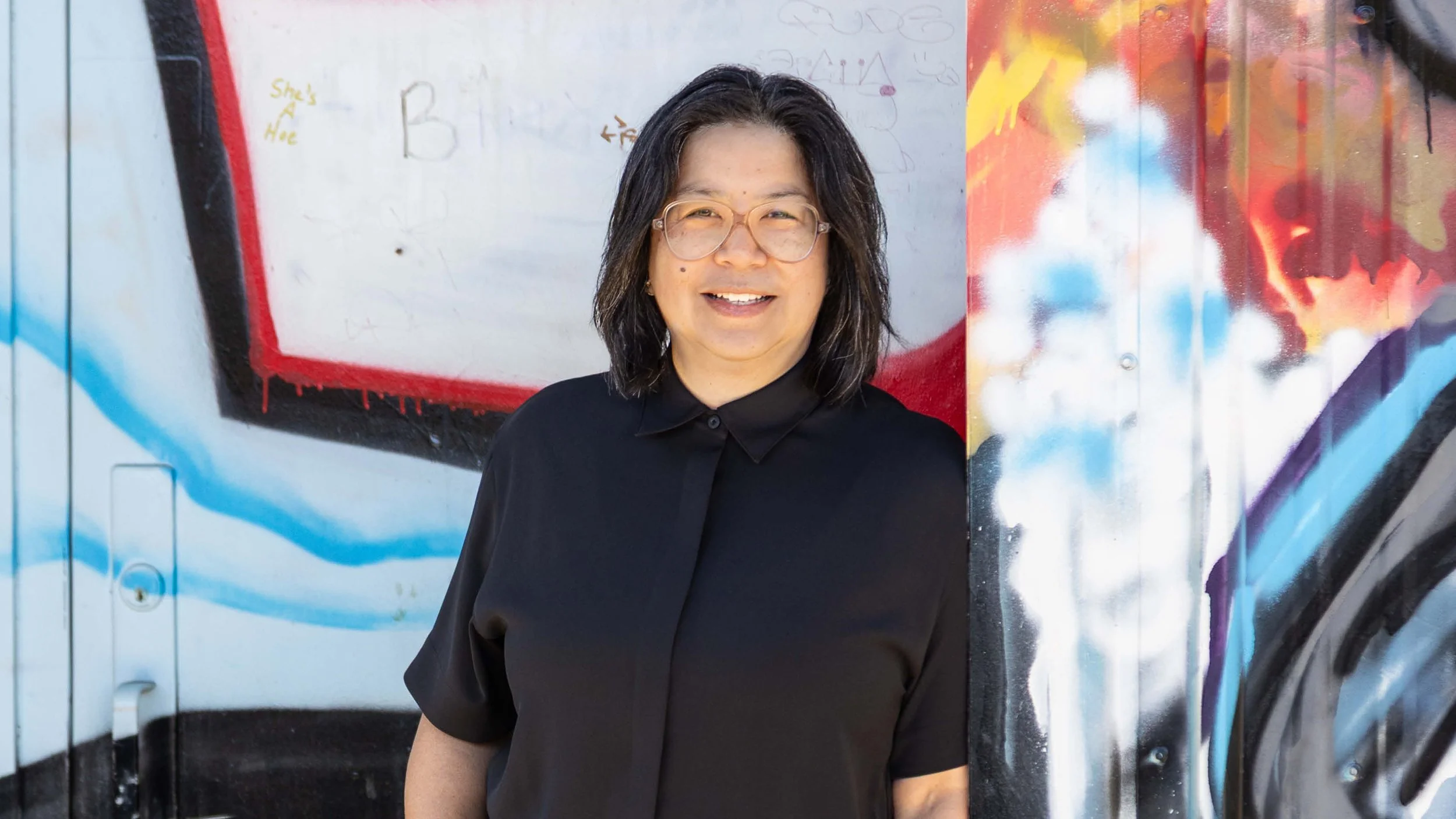 An image of a smiling person in a black button up top in front of a brightly painted exterior wall.