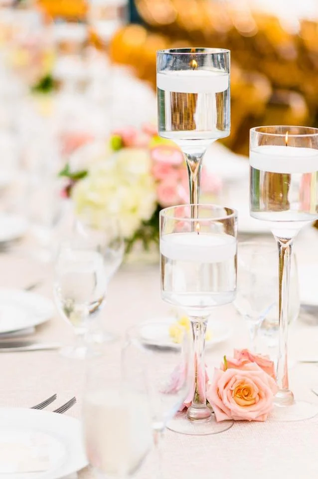 Elegant table setting with tall glass candle holders filled with water and floating candles, pink roses, and soft, blurred background with warm lighting.