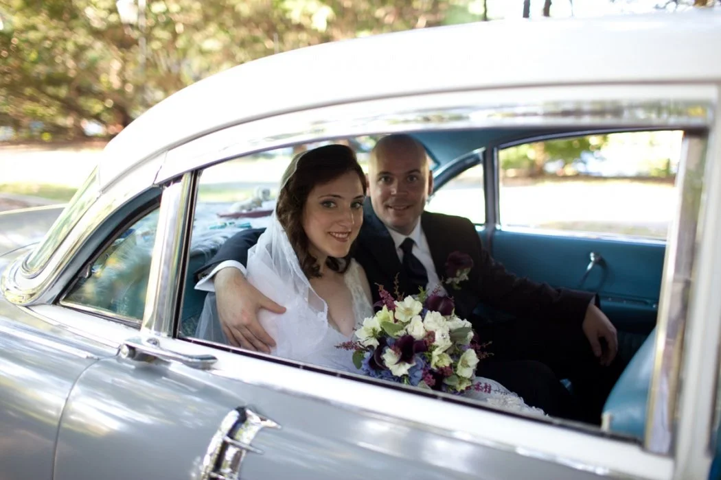 A bride and groom sitting in the backseat of a vintage car, smiling. The bride holds a bouquet of white and purple flowers. The groom is dressed in a black suit and tie.