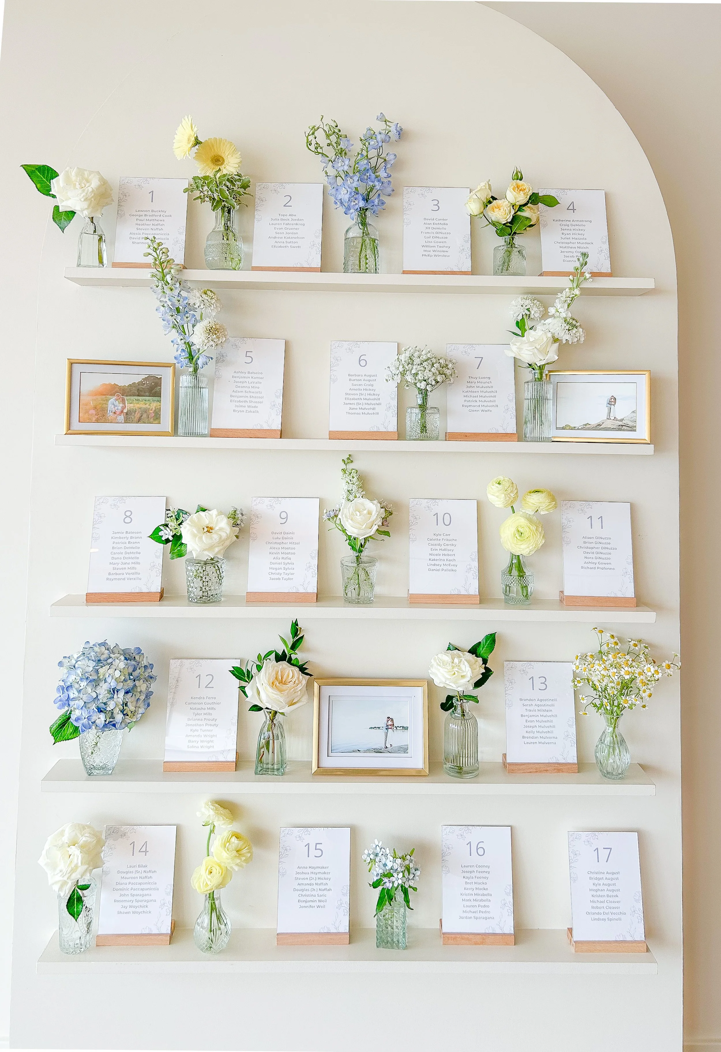 Decorative wall display featuring three white shelves with small vases containing white, yellow, and blue flowers, along with framed photographs and seating cards with names.