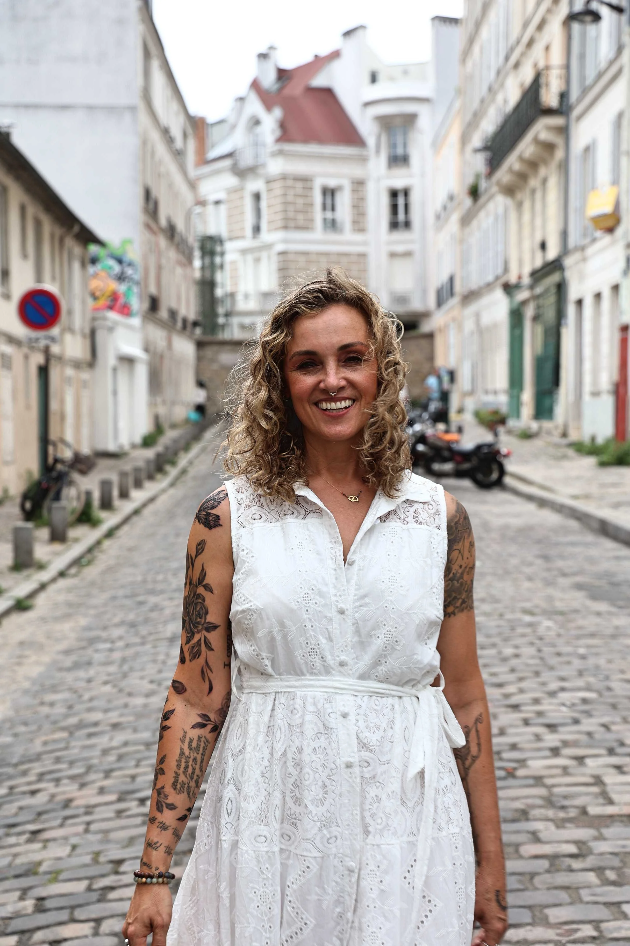 Smiling woman with curly hair and tattoos, wearing a white dress, stands on a cobblestone street in a European city.
