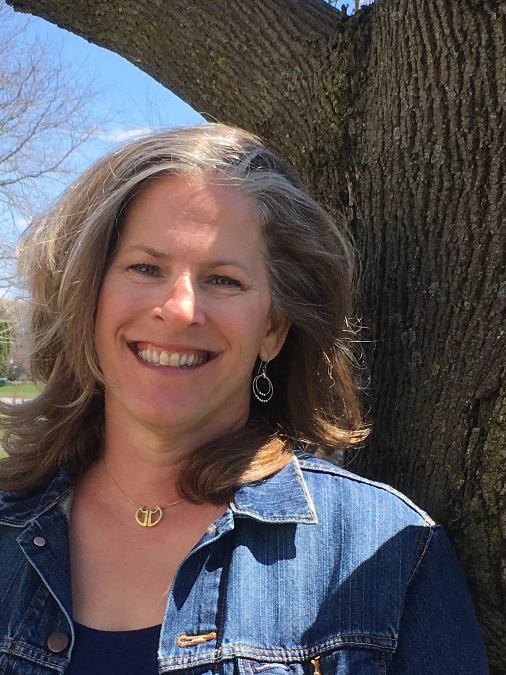 Woman smiling in front of a tree wearing a denim jacket and necklace outdoors on a sunny day.