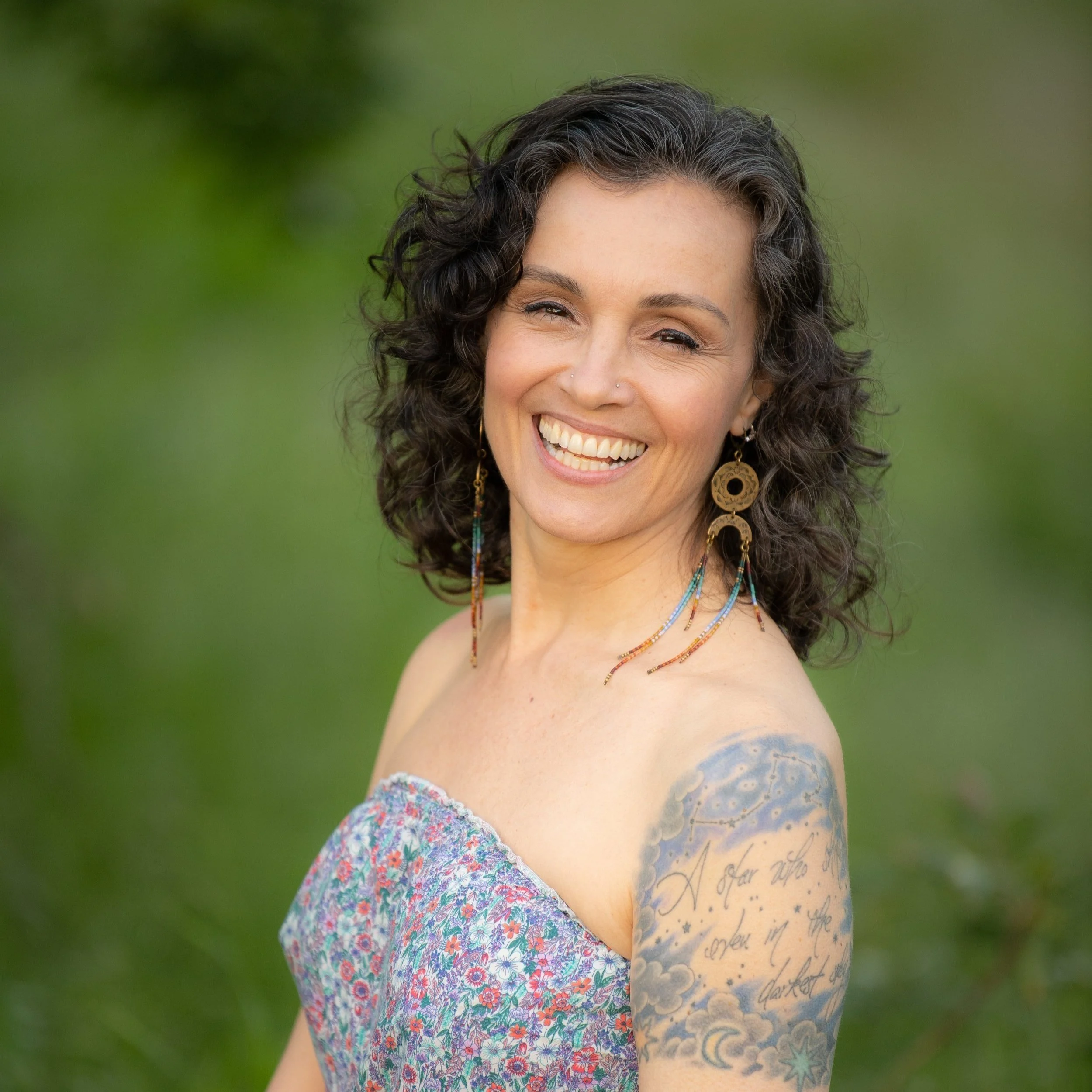 Smiling woman with curly hair, wearing large earrings and a floral dress, showing a tattoo on her arm.