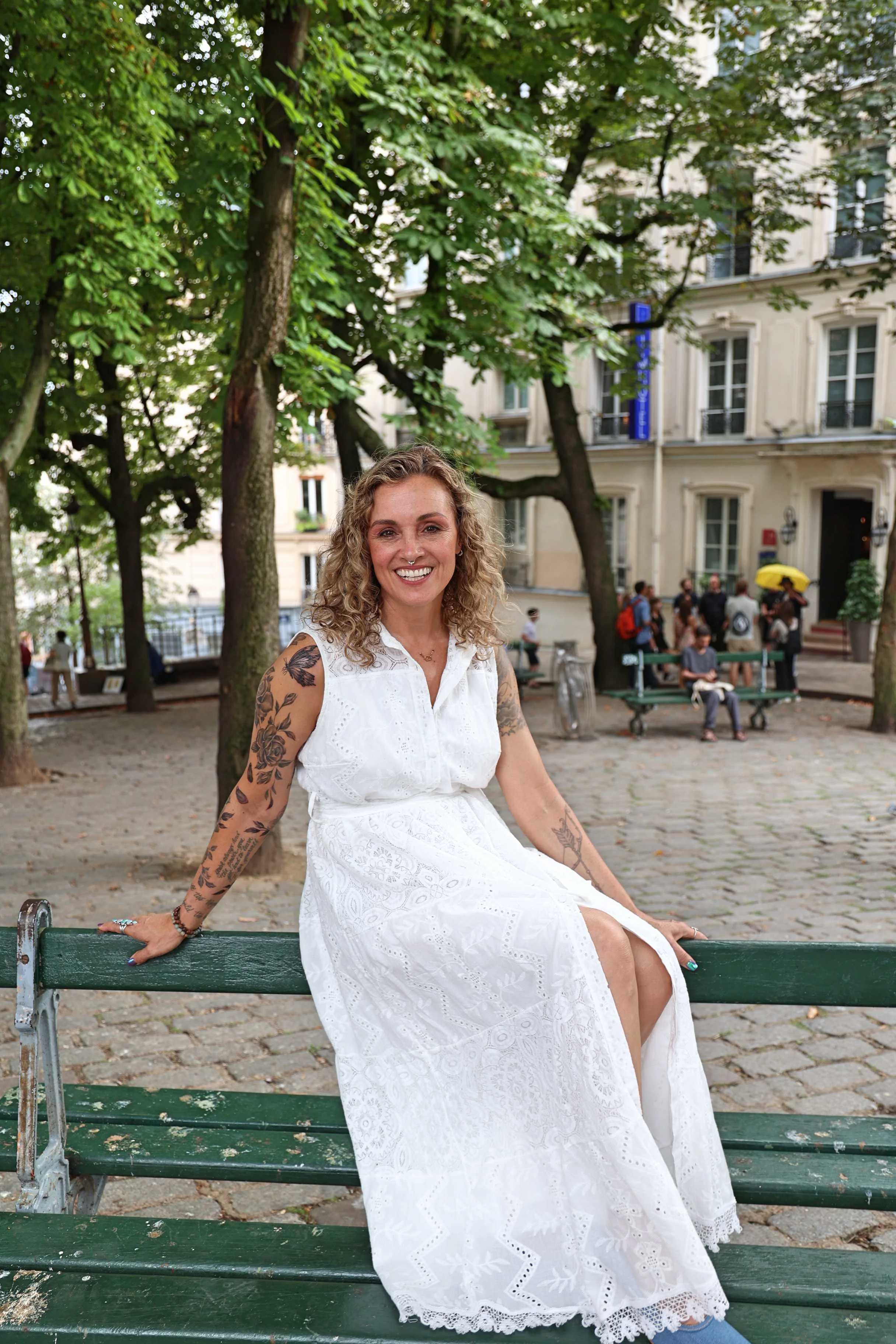 Smiling woman with tattoos in white dress sitting on park bench under trees, urban background