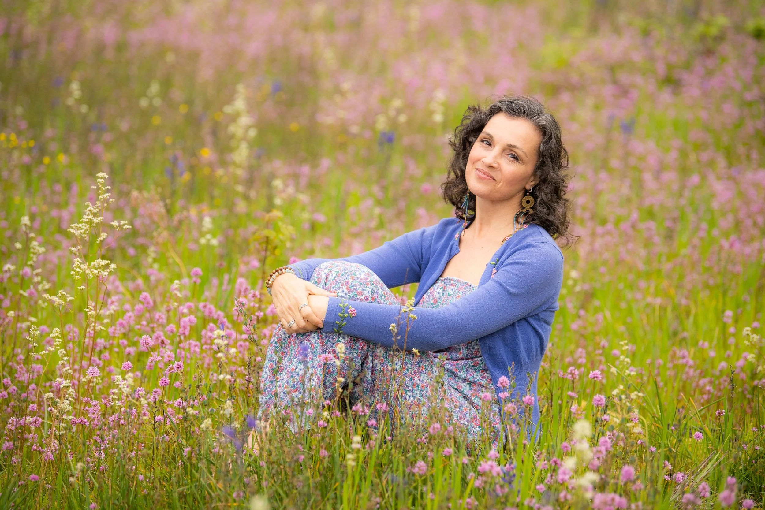 Woman sitting in a field of wildflowers, wearing a blue cardigan and floral dress.