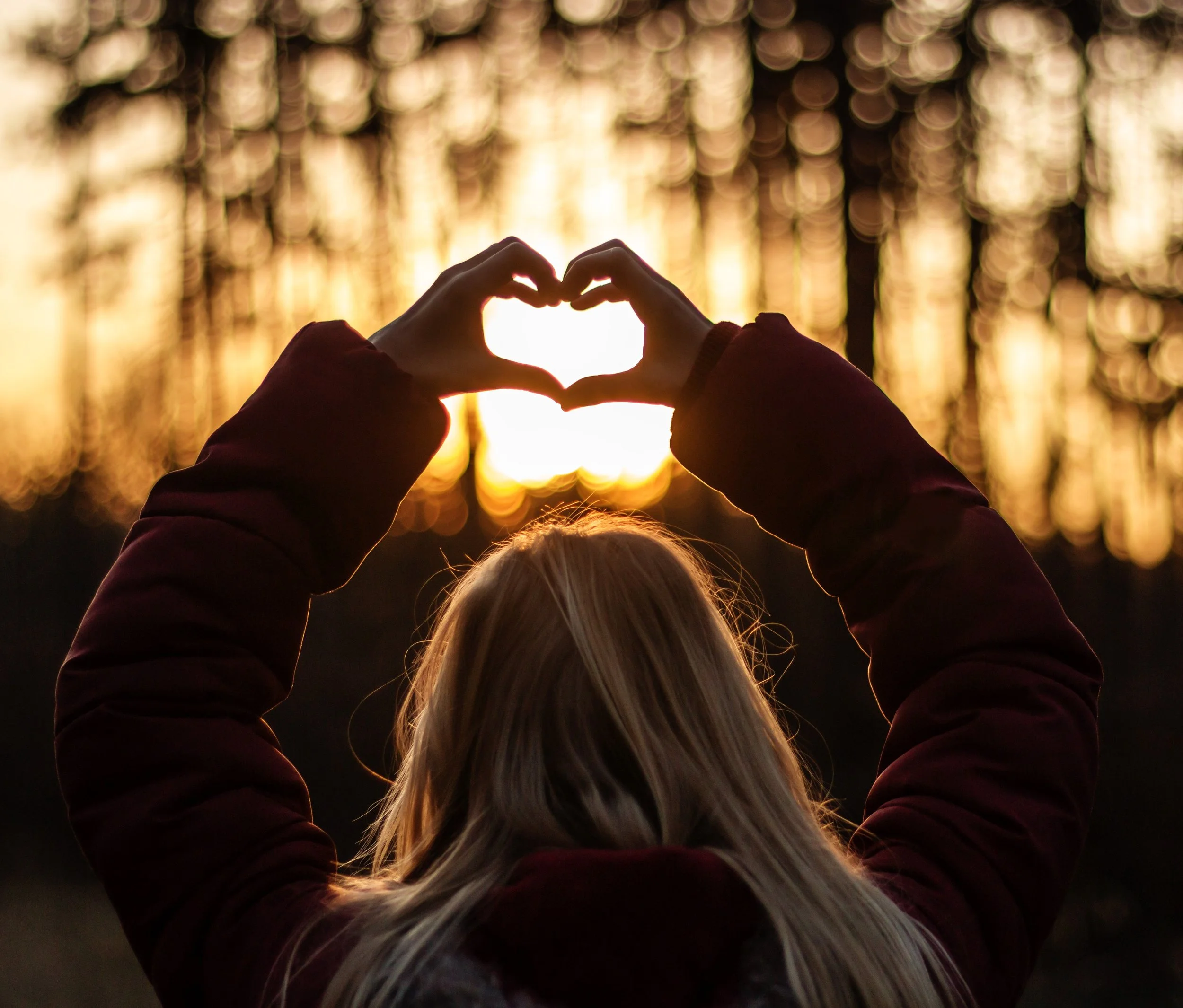 Silhouette of a person forming a heart shape with their hands against a sunset backdrop in a forest.