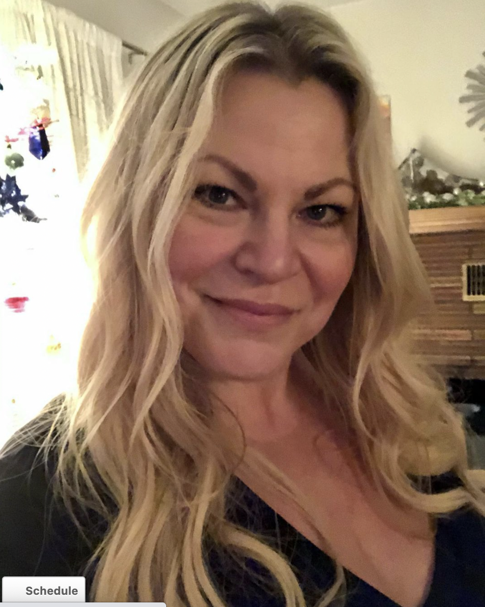 Blonde woman smiling indoors with long hair, wearing a black top, and standing near a softly lit background.
