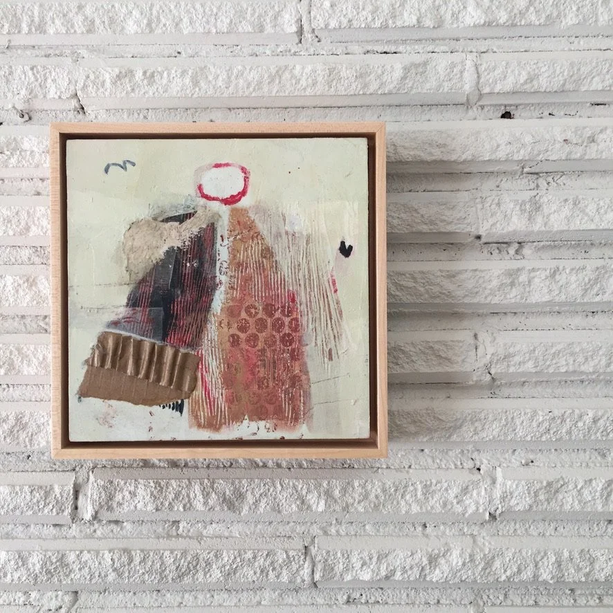 Abstract mixed-media artwork in a wooden frame hanging on a white brick wall, featuring various textures, shapes, and colors including red, beige, black, and white.