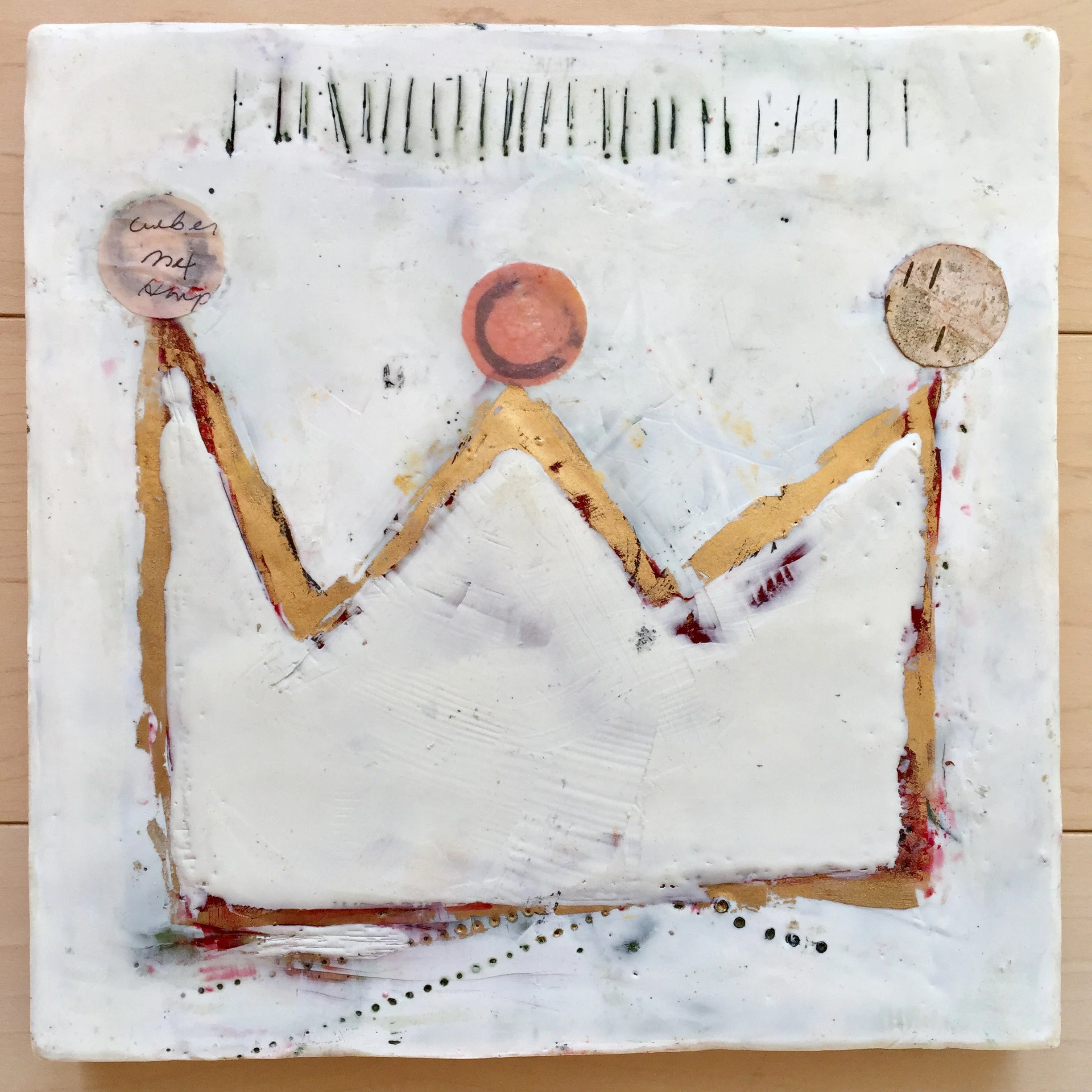 A white art piece with three round, textured elements connected by curved, orange and yellow lines, featuring black marks and dotted lines, placed on a light wooden surface.