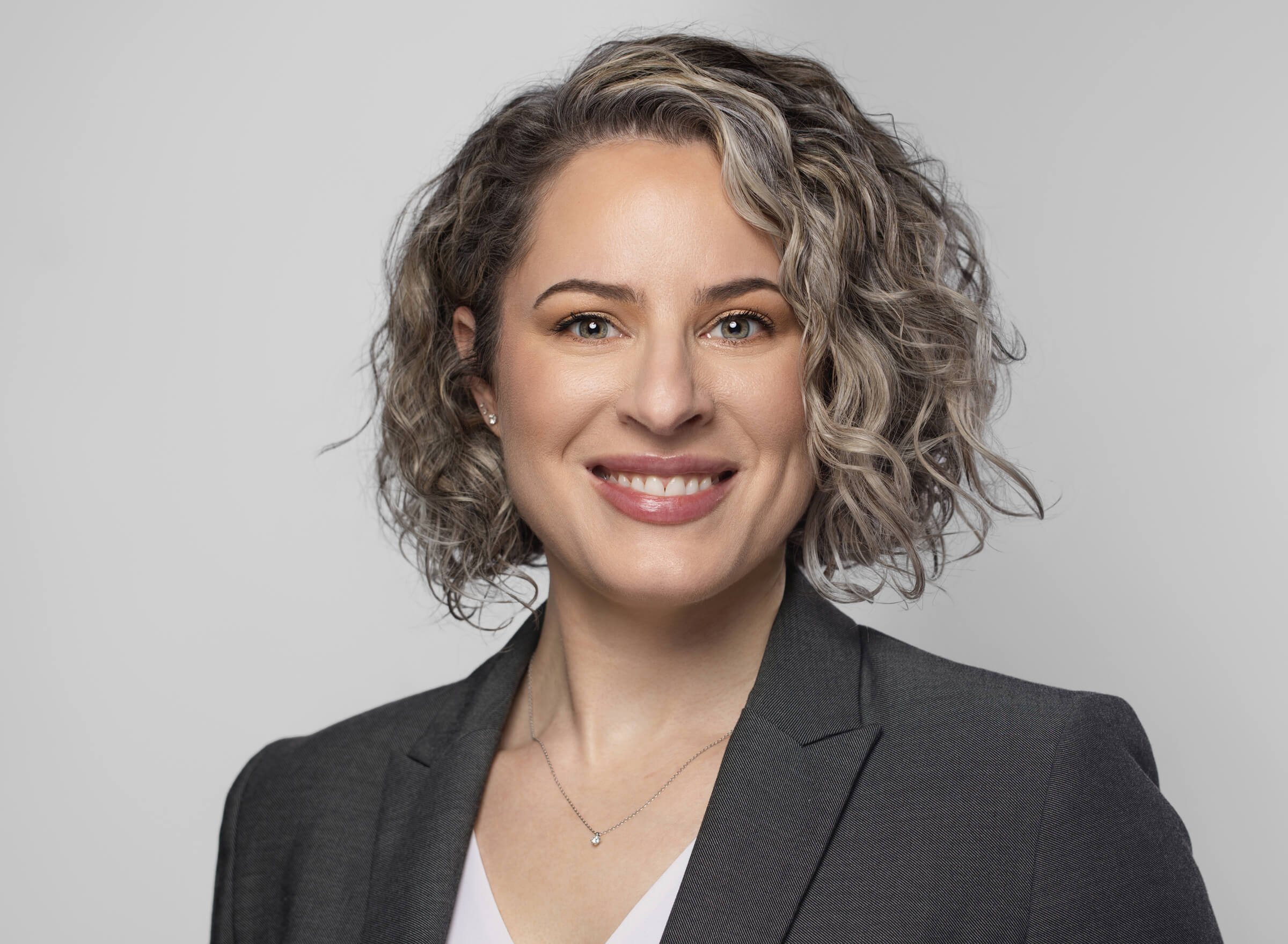Smiling person with curly hair in a gray blazer, neutral background.