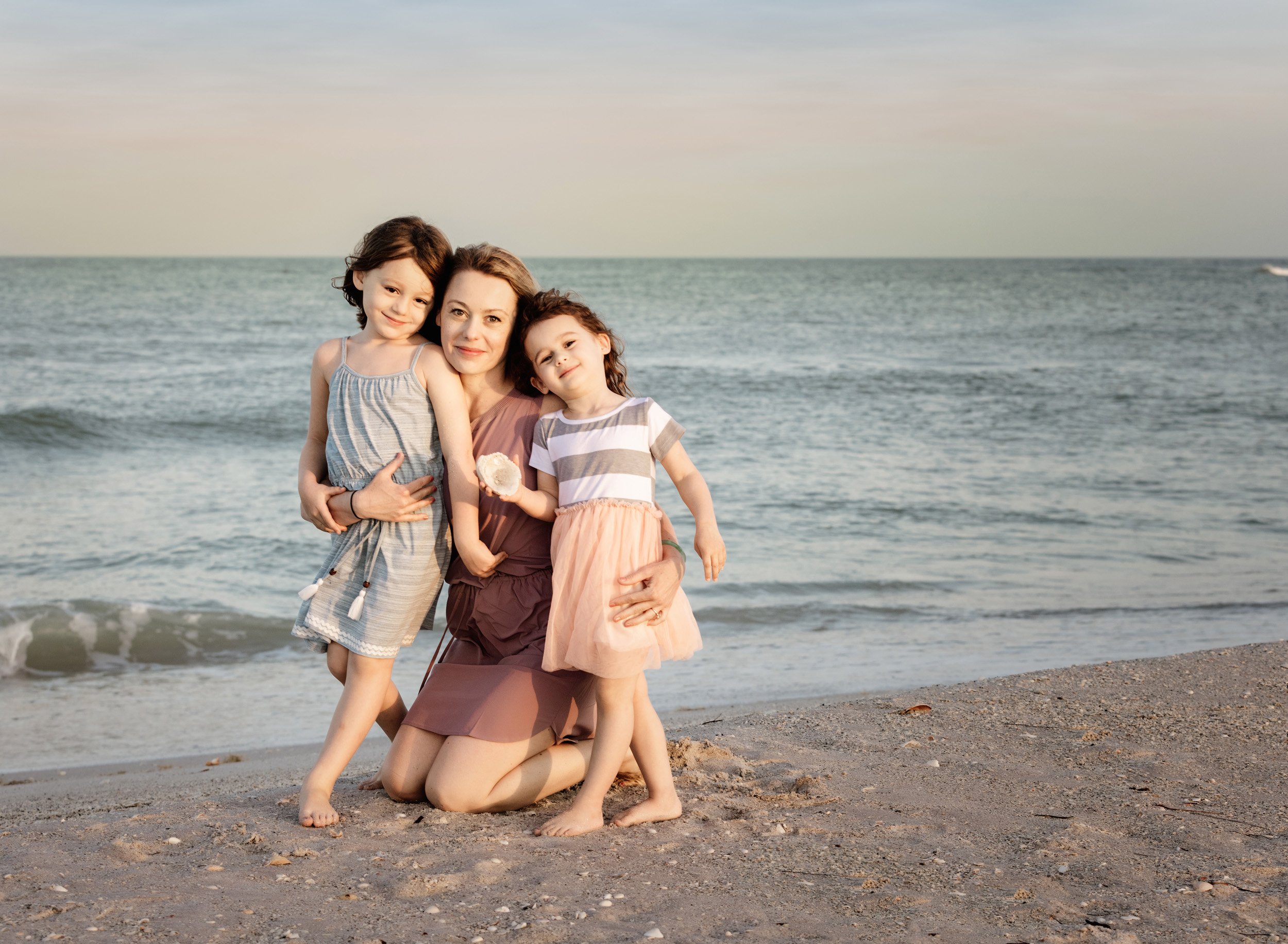A woman kneeling on a beach with two young girls, smiling at the camera. One girl is holding a seashell. The ocean is in the background.