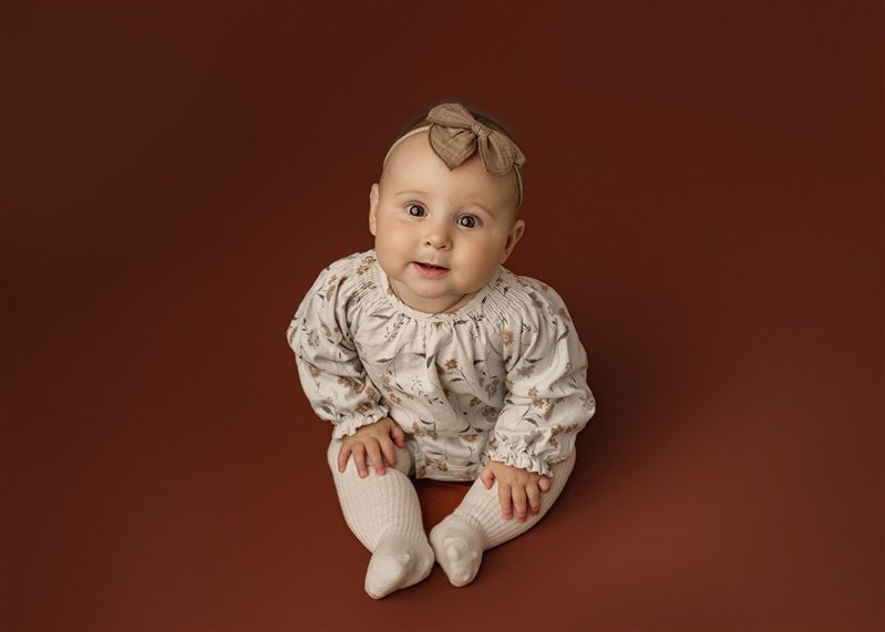 A baby wearing a floral-patterned outfit with a headband bow, sitting against a brown background.