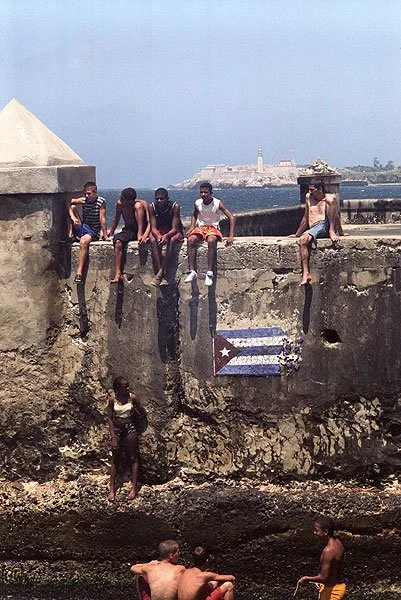 Children sitting and standing along a historic stone wall with a mural of the Cuban flag painted on it, overlooking the sea with forts and a lighthouse in the background.