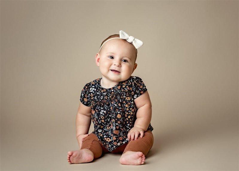 Baby wearing a floral top and brown leggings, sitting and smiling with a white bow headband on.