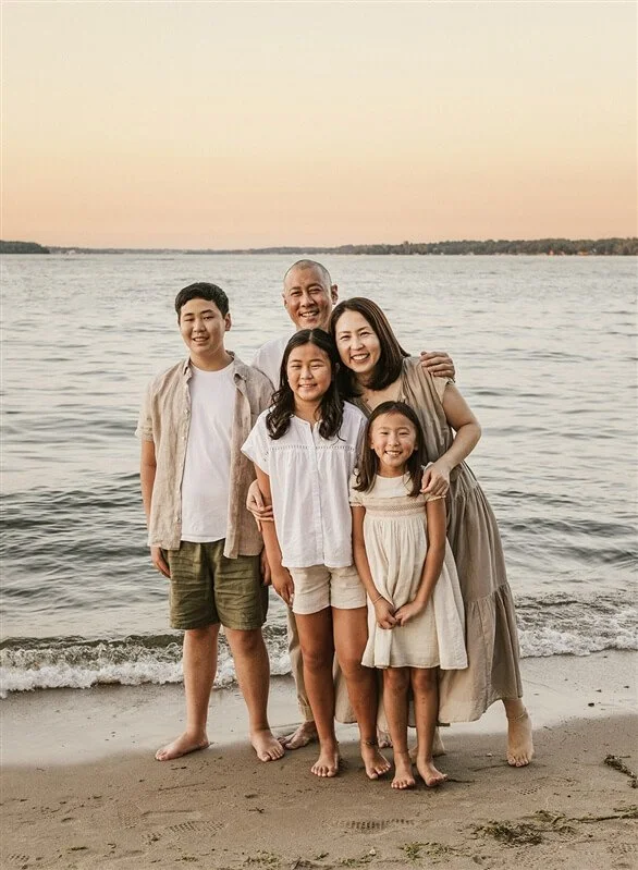 Family of five standing on the beach at sunset, smiling and embracing, near the water with sandy shore and distant land in the background.