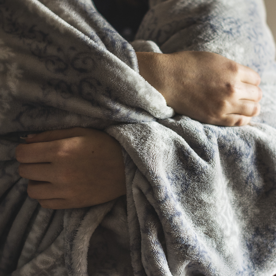 A person wrapped in a cozy blanket, sitting, with only their hands and part of torso visible, conveying a sense of warmth and safety.