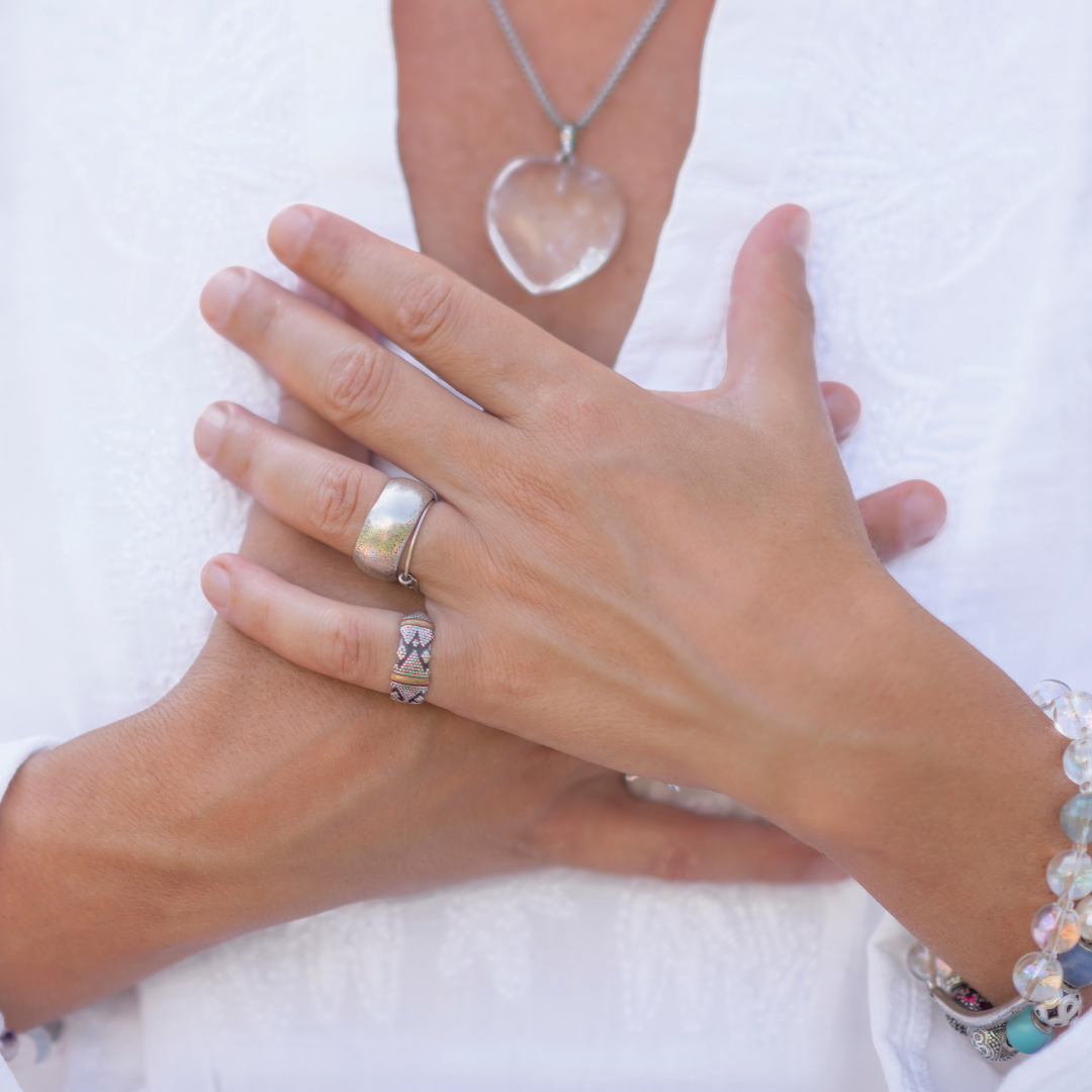 A woman in white clothes and a heart-shaped crystal necklace, hands over her heart, conveying self-love.