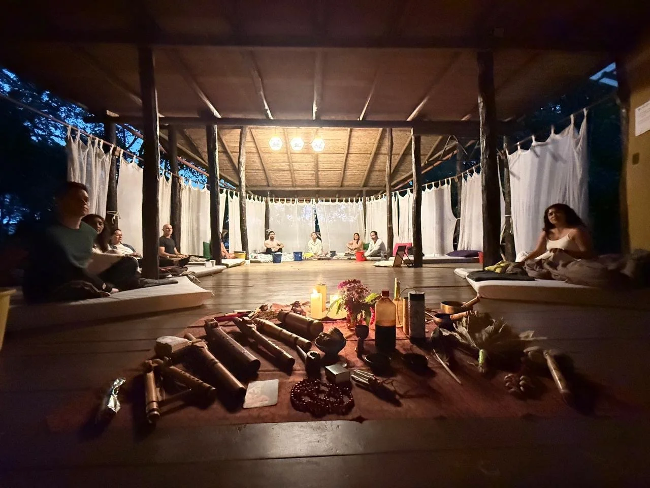 Reunión en un espacio abierto con personas sentado en cojines, en el centro objetos tradicionales y velas.