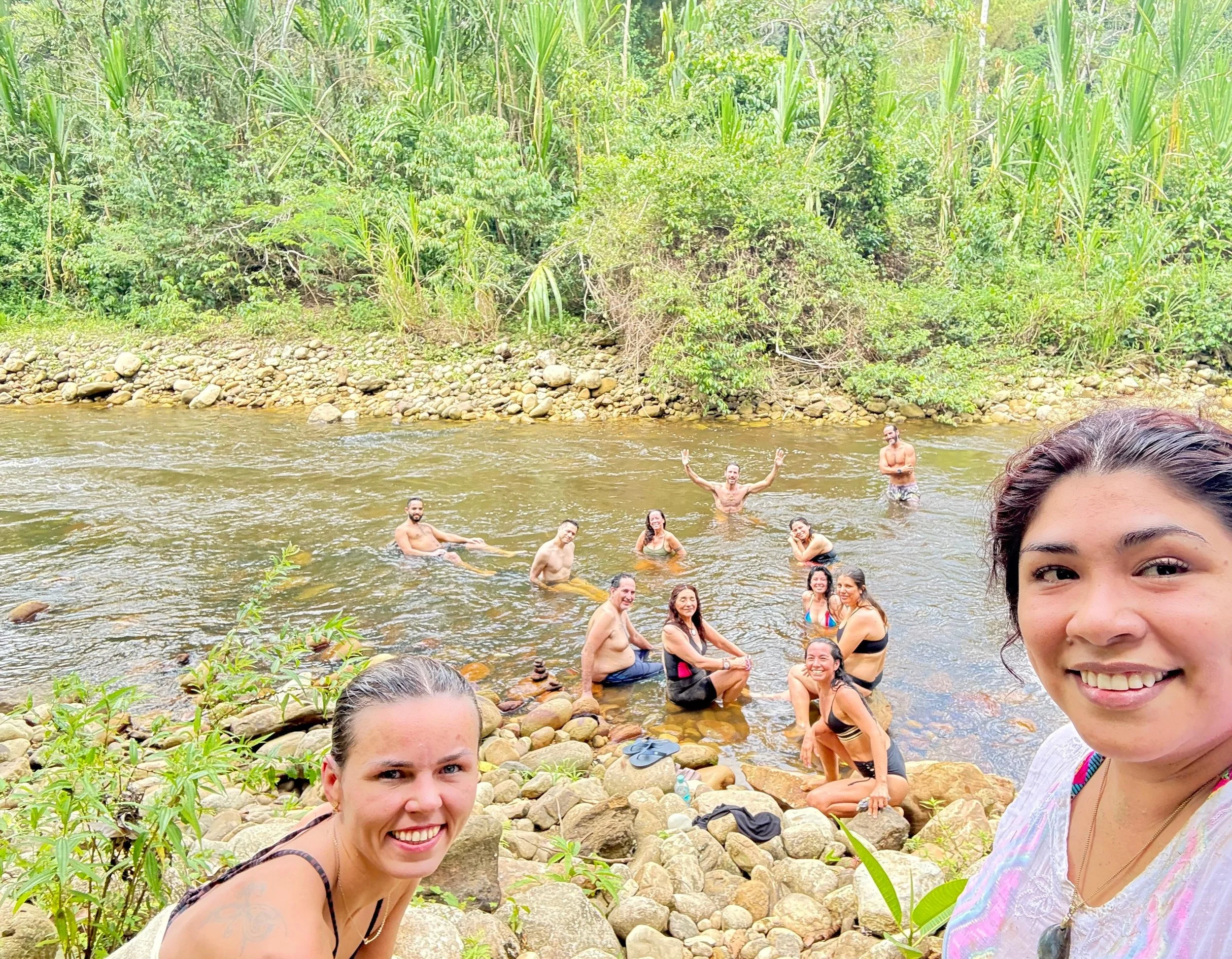 Grupo de personas disfrutando en una río rodeado de vegetación tropical, algunas están en el agua y otras en las piedras en la orilla, todos se ven felices y relajados.