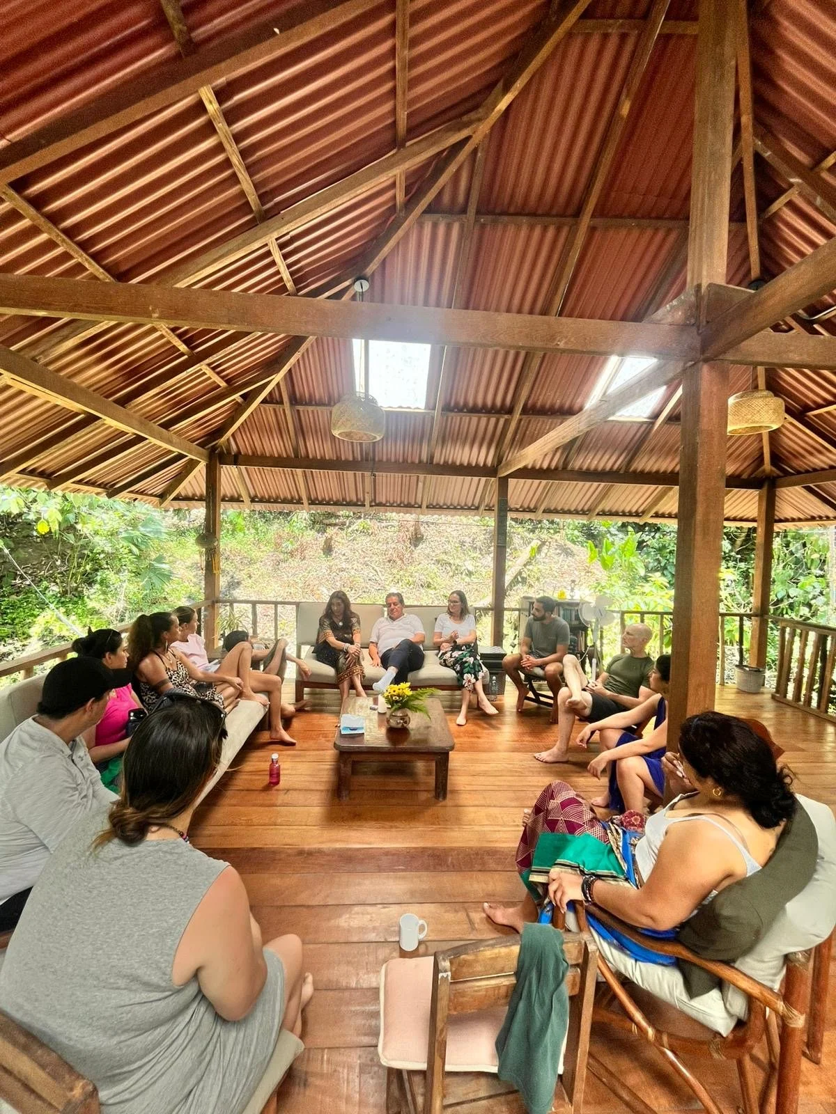 Un grupo de personas sentado en un salón de madera, con techo de tejas, en una reunión o conversación en un entorno natural y tranquilo.