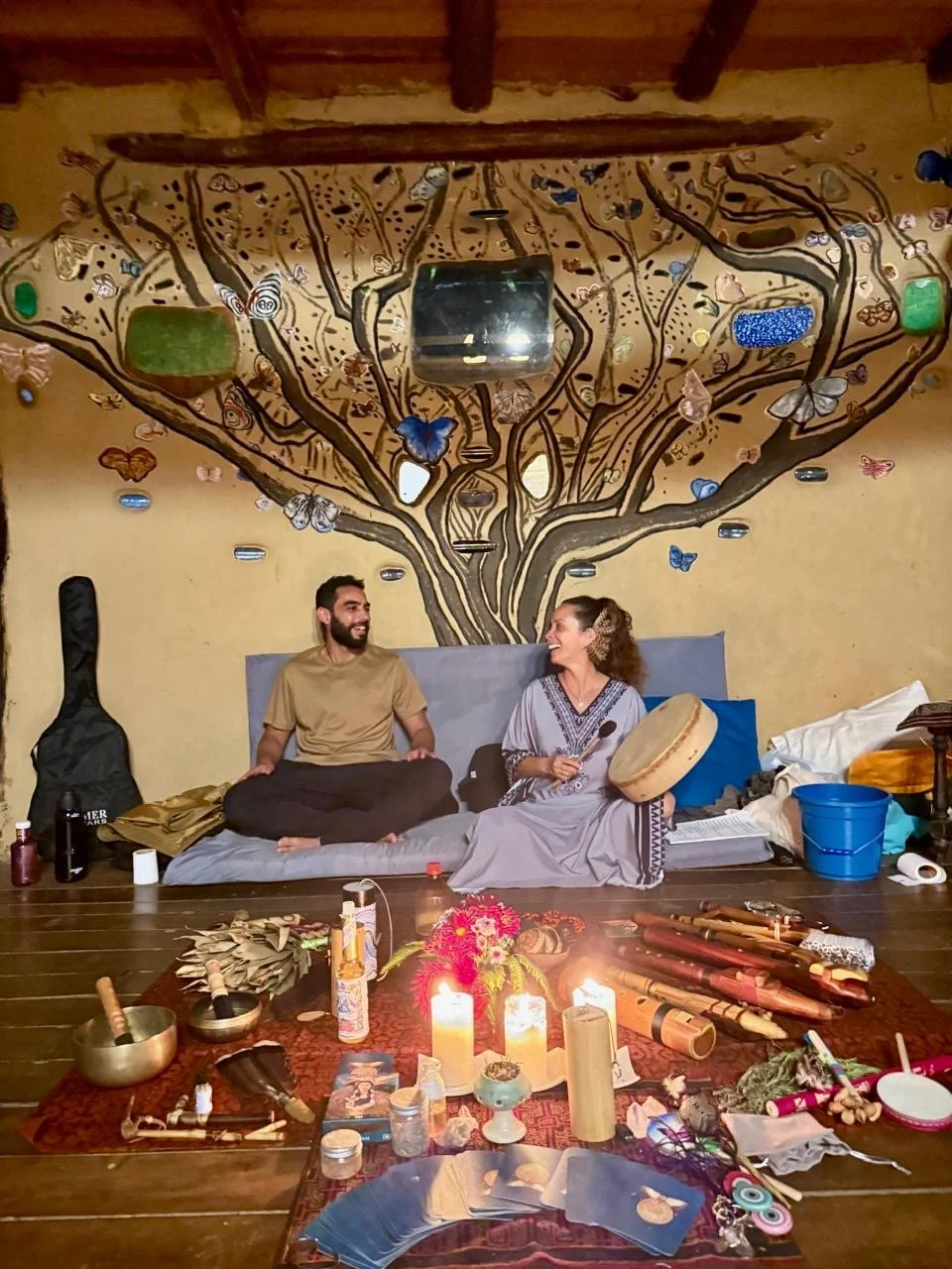 Una pareja está sentado en el suelo en una habitación decorada con un mural de árbol en la pared, rodeados de instrumentos musicales y objetos rituales, con velas encendidas en una mesa frente a ellos.