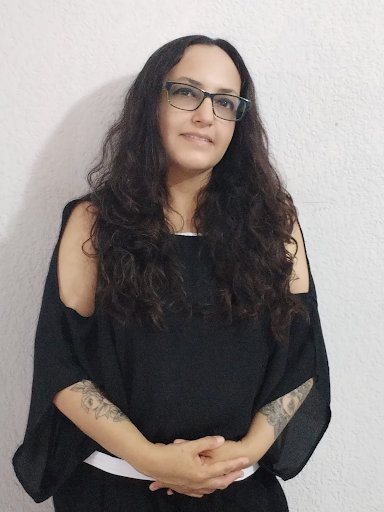 Mujer con cabello rizado y oscuro, usa gafas y una blusa negra con los hombros descubiertos, frente a una pared blanca.